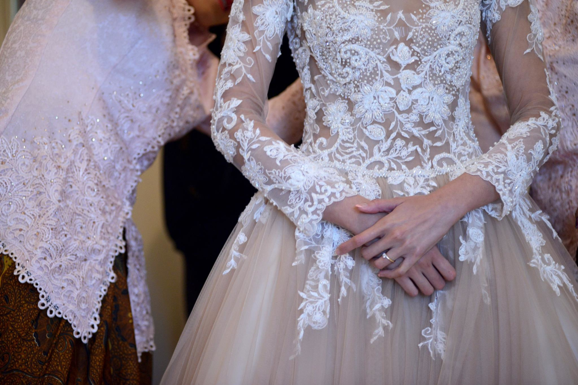 Bride in an intricate lace long-sleeve wedding gown being assisted while getting ready.