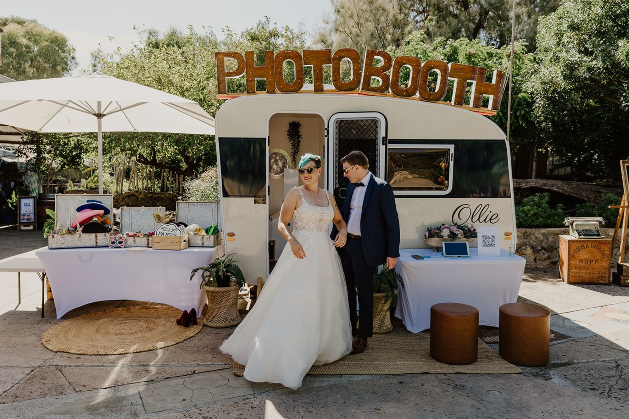 Newlywed couple poses in front of a retro caravan photobooth setup at an outdoor wedding.
