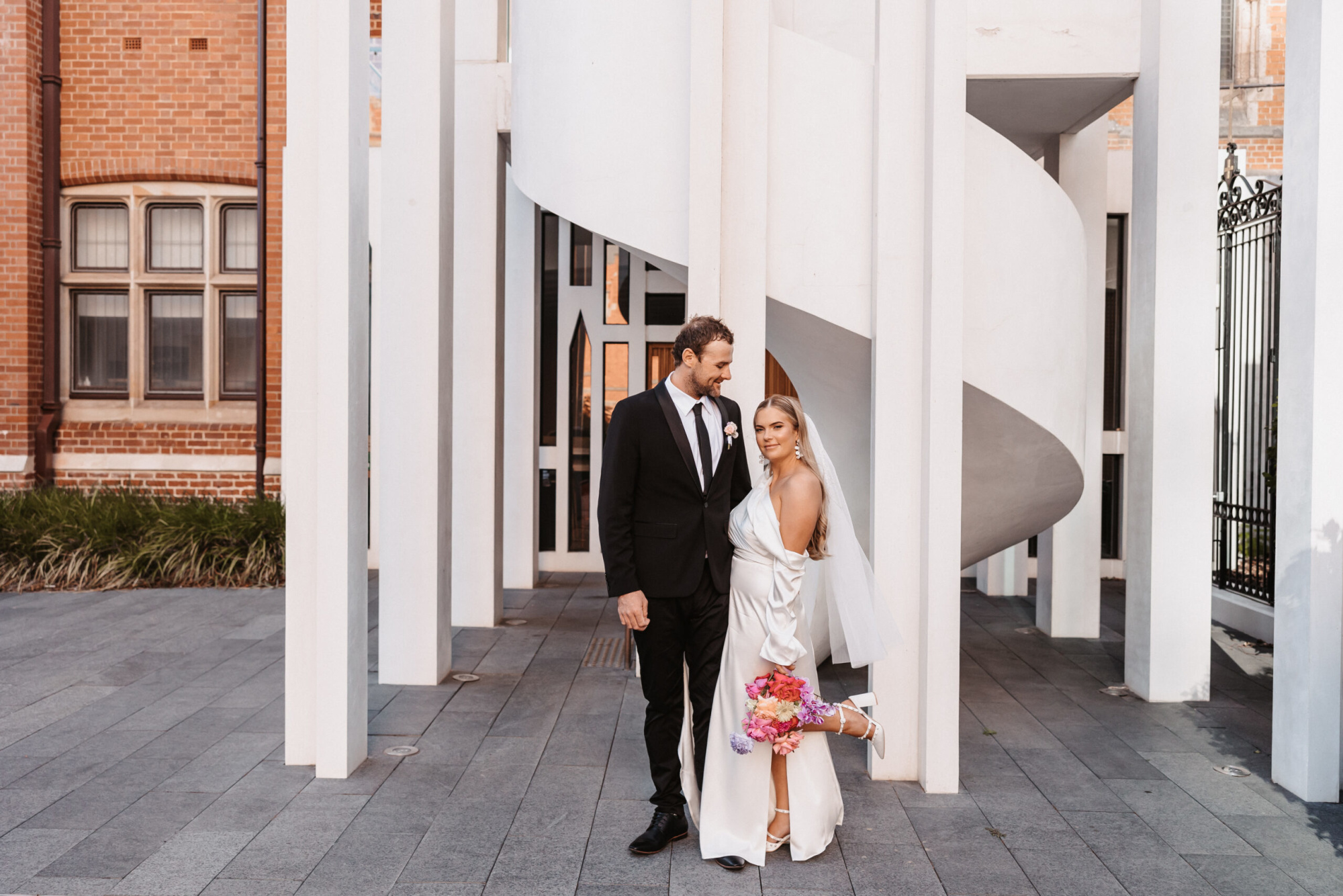 Bride and groom pose in front of modern white architectural columns with a spiral staircase and brick building backdrop.