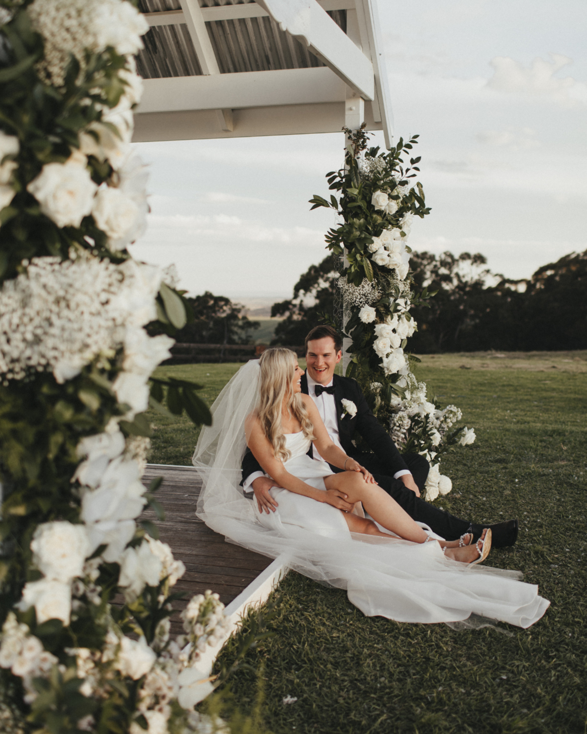 Bride and groom sit together beneath a flower-covered outdoor gazebo at a garden wedding.