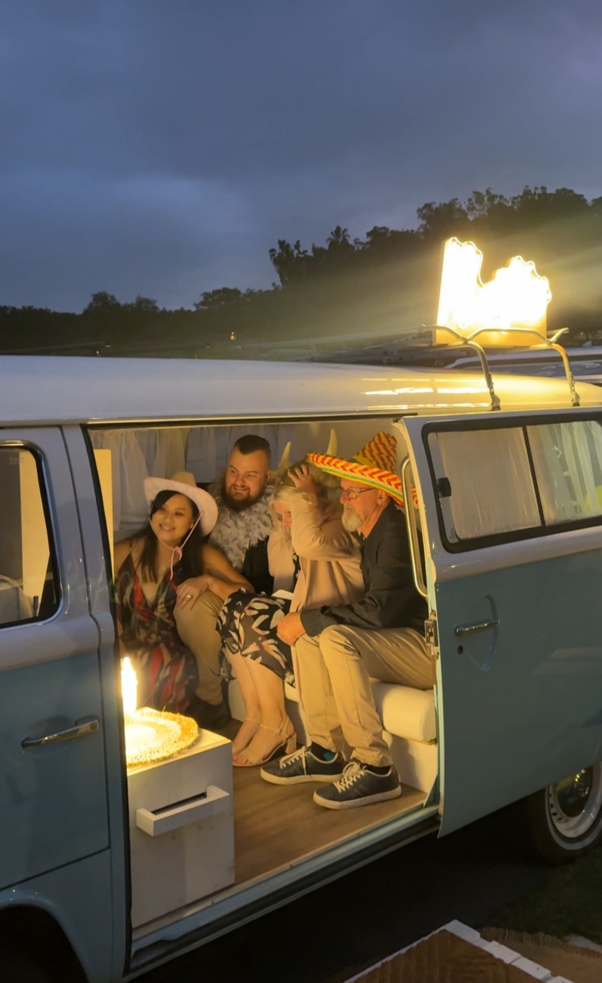 Wedding guests wearing fun hats pose inside a vintage camper van photo booth at night.