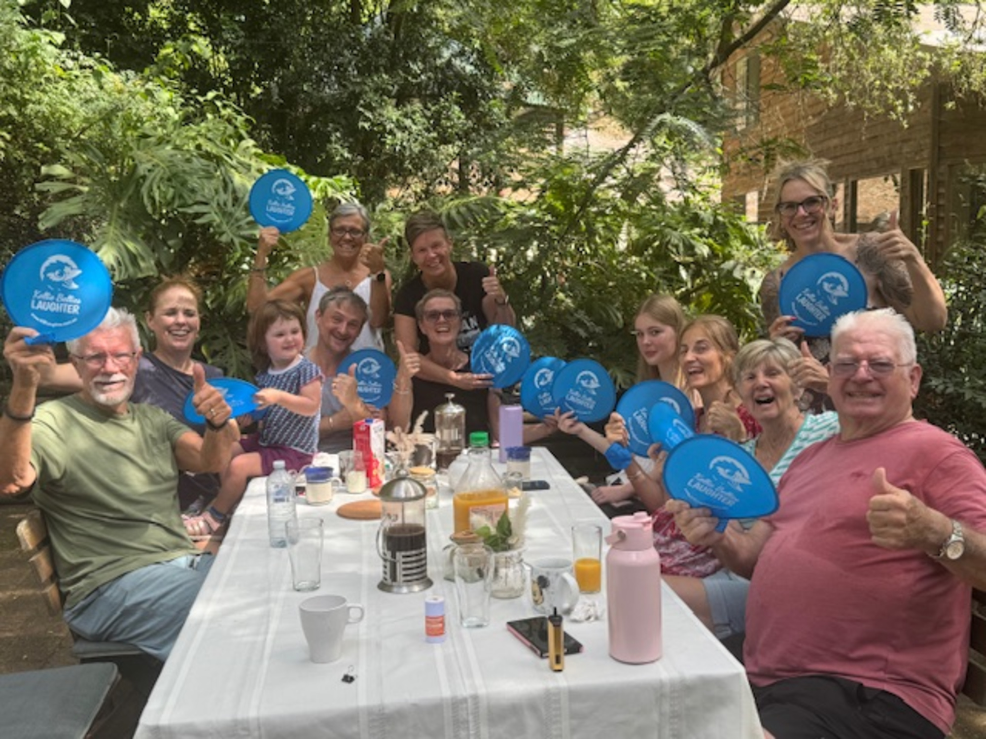 A large group of people smile and pose with blue fans around a long table at an outdoor garden gathering.
