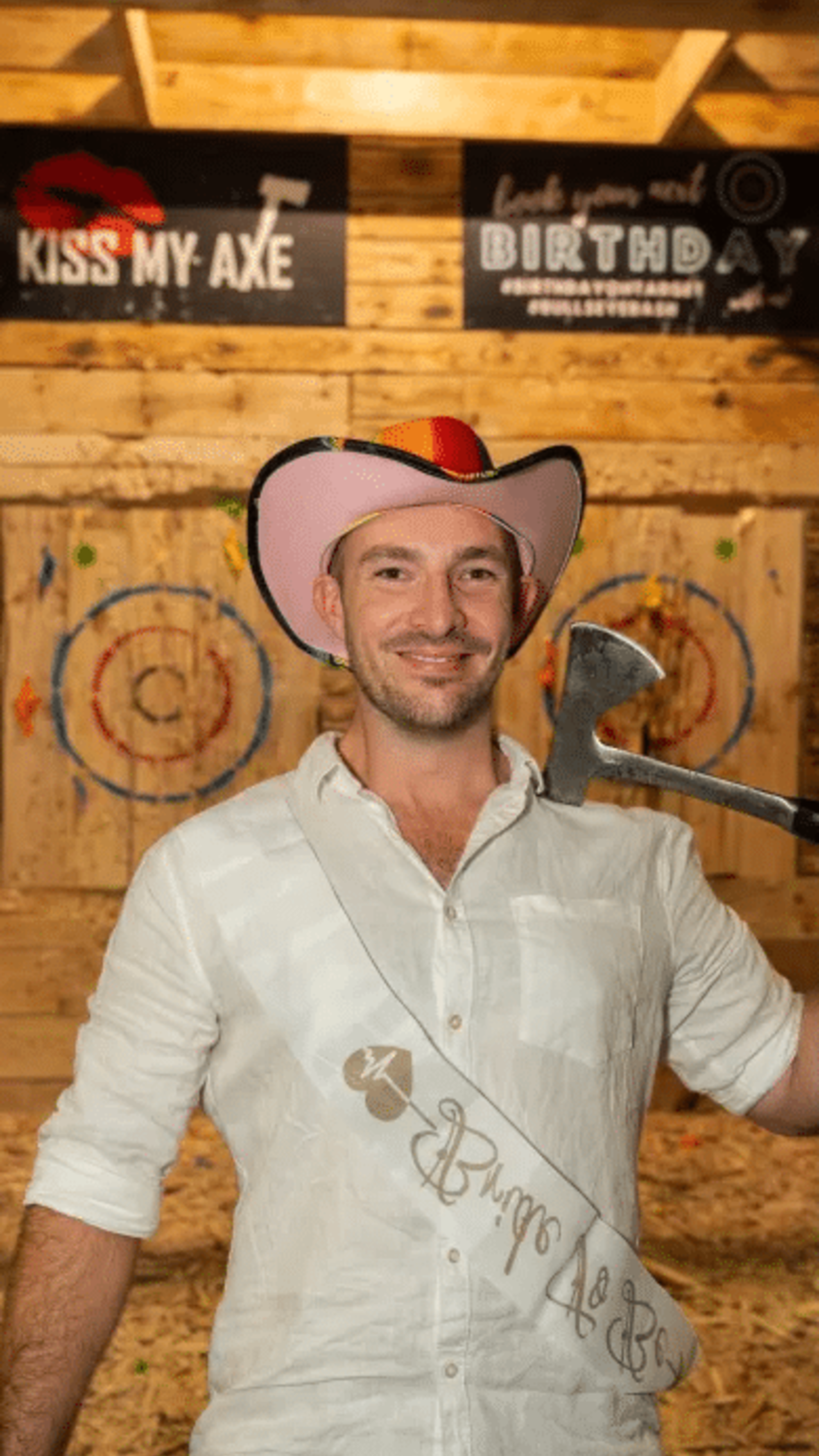 Smiling groom-to-be in a pink cowboy hat holding an axe at an indoor axe-throwing party venue.