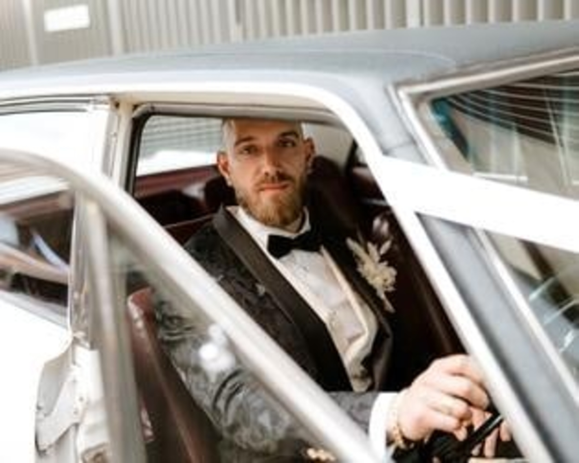 Groom in a black tuxedo sitting inside a classic white wedding car looking toward the camera.