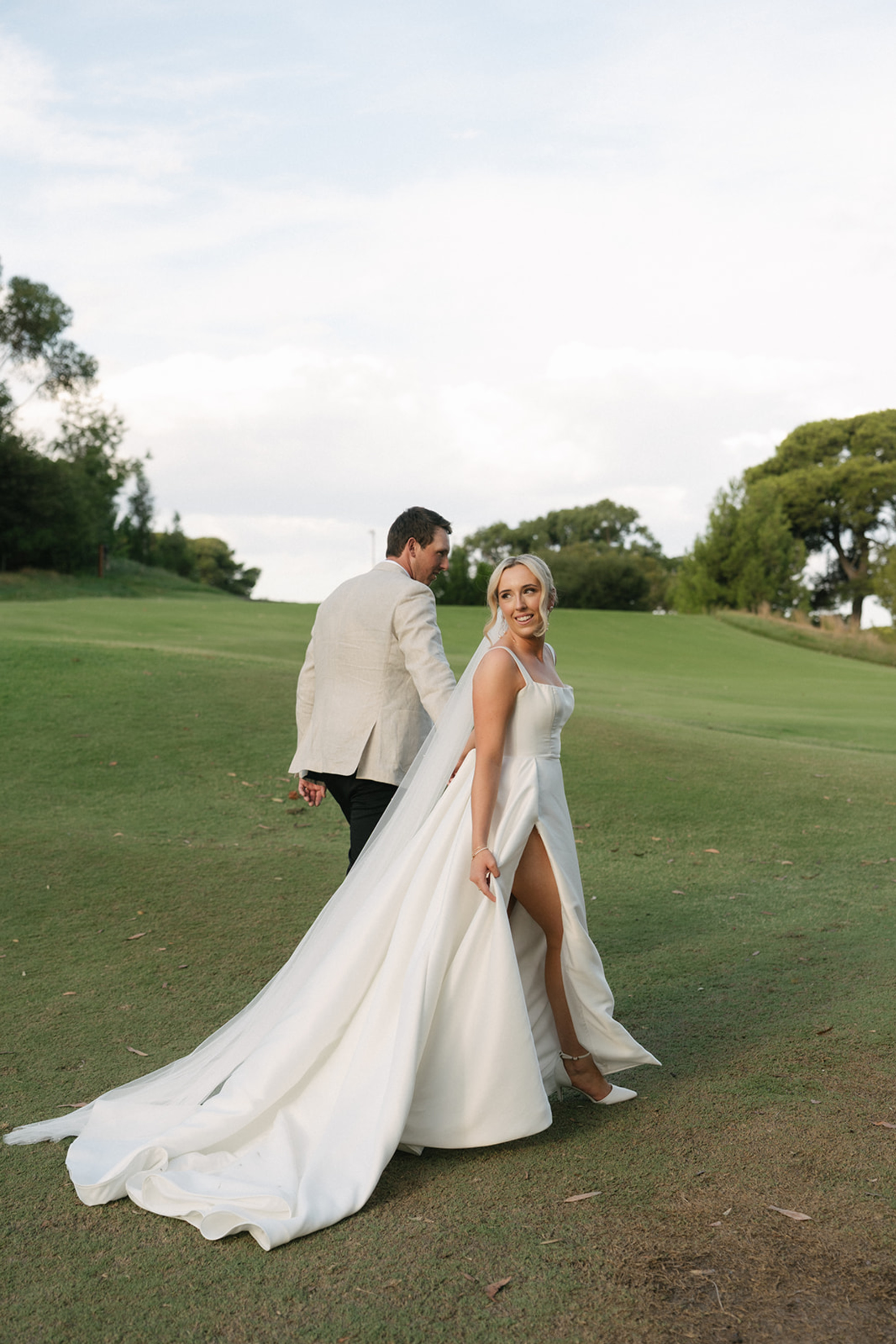 Bride and groom walk across a green lawn, the bride in a flowing white gown with a slit and long train.