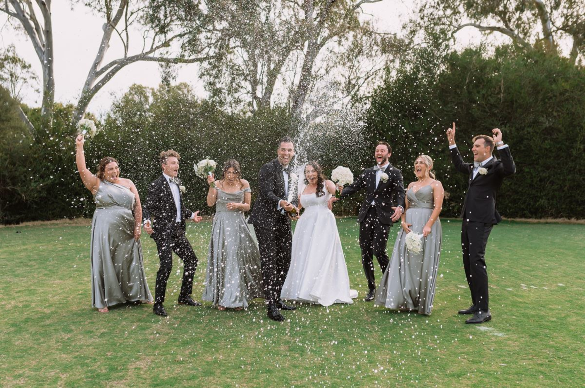 Bride and groom pop champagne while celebrating with their wedding party on a green lawn.