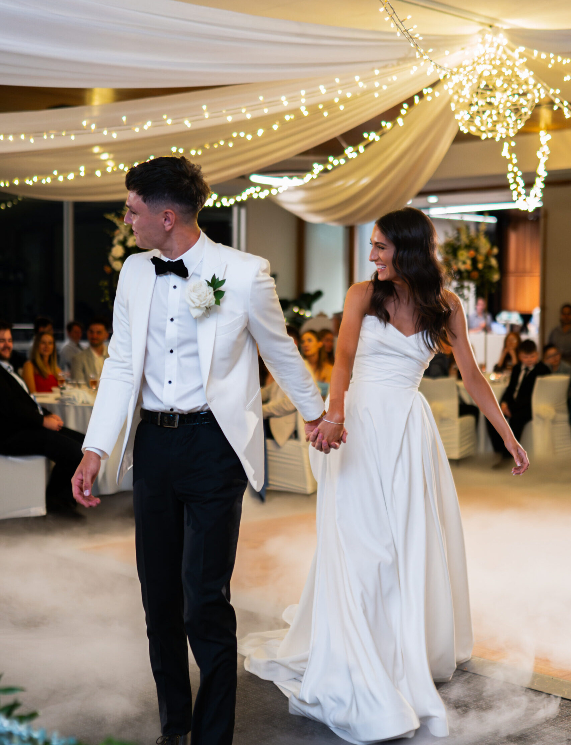 Bride and groom hold hands walking through a fog-filled, fairy-light decorated wedding reception.