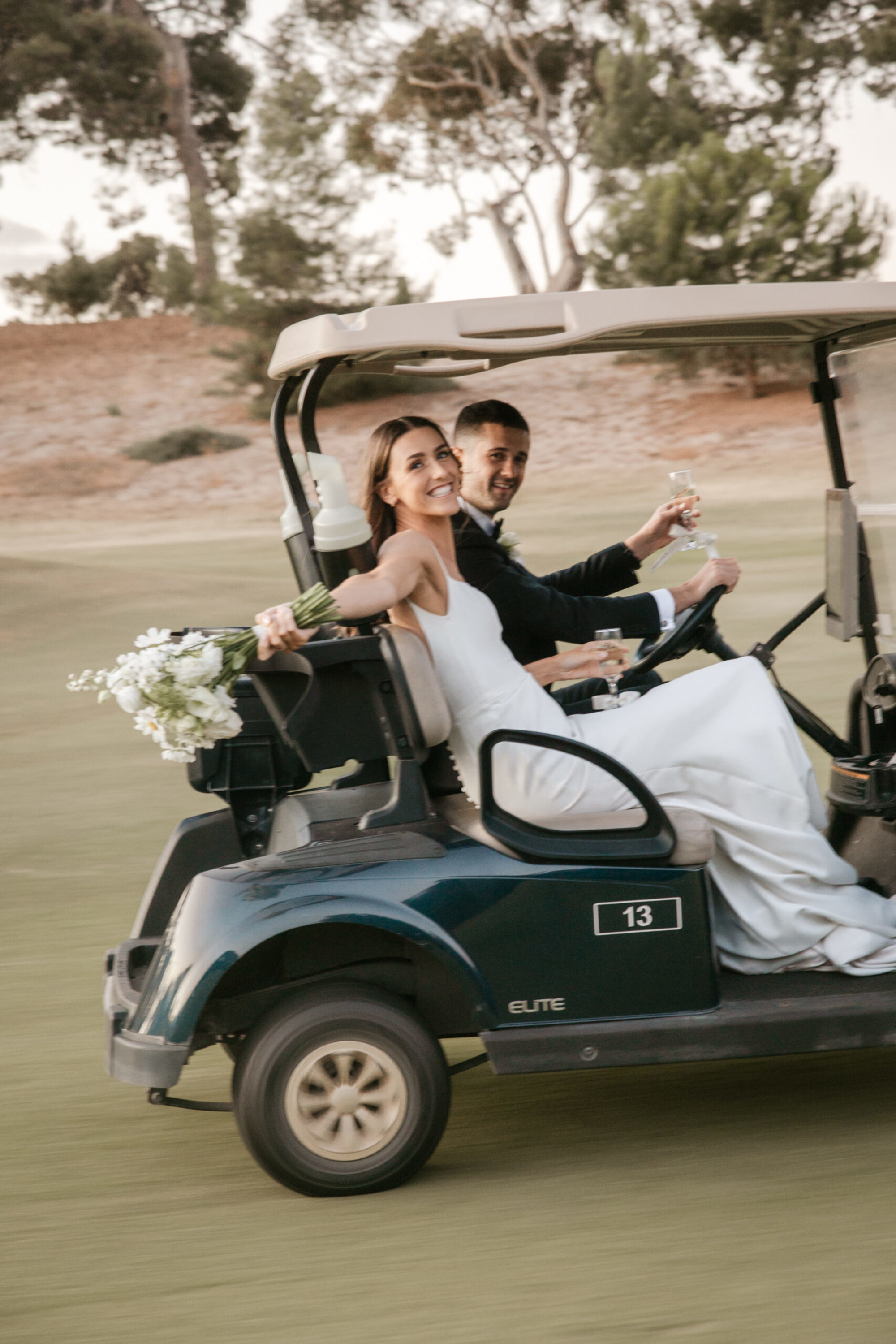 Bride and groom ride a golf cart across a golf course, smiling and holding champagne and a white bouquet.