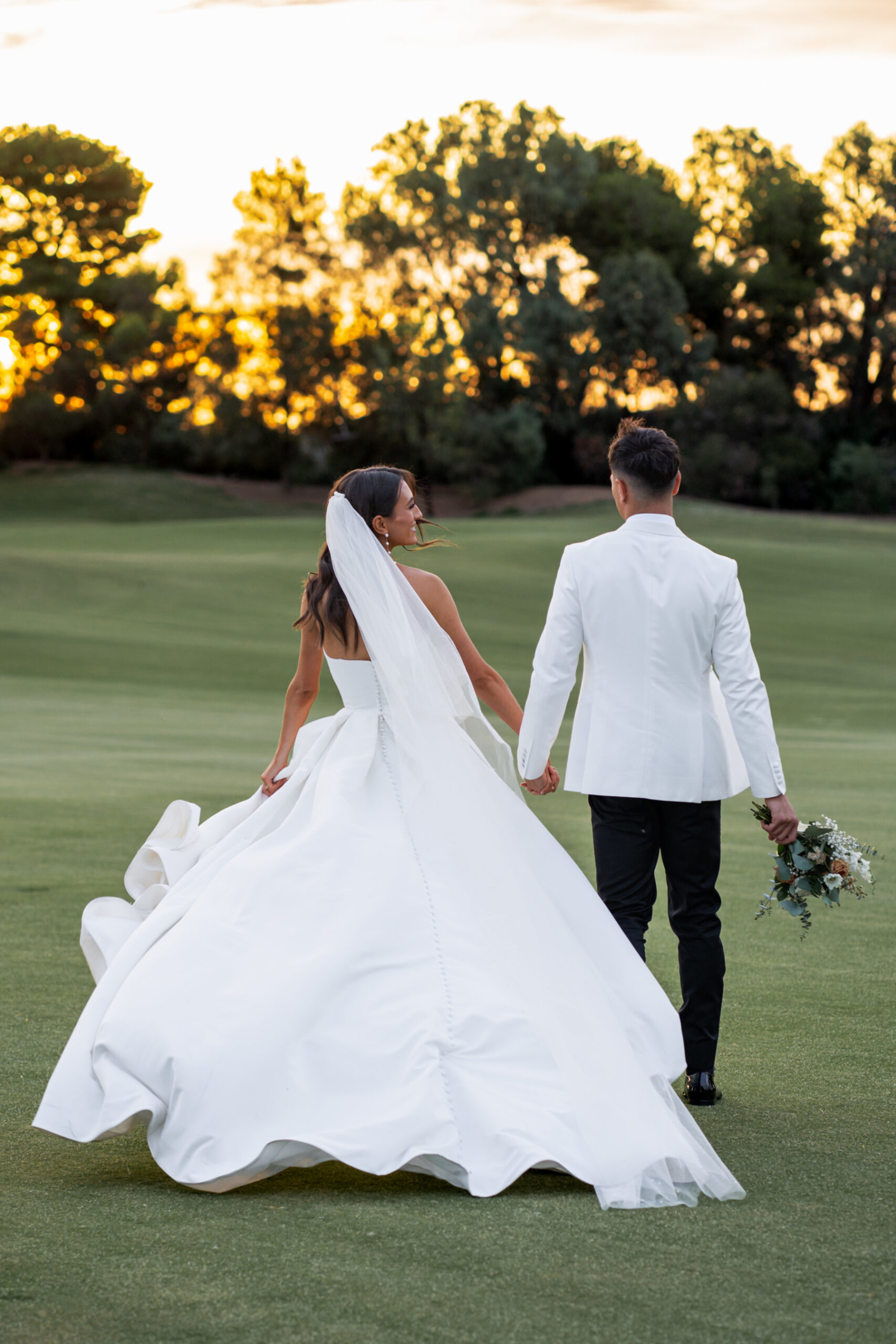 Bride and groom hold hands walking across a golf course at sunset, her full white gown flowing behind them.