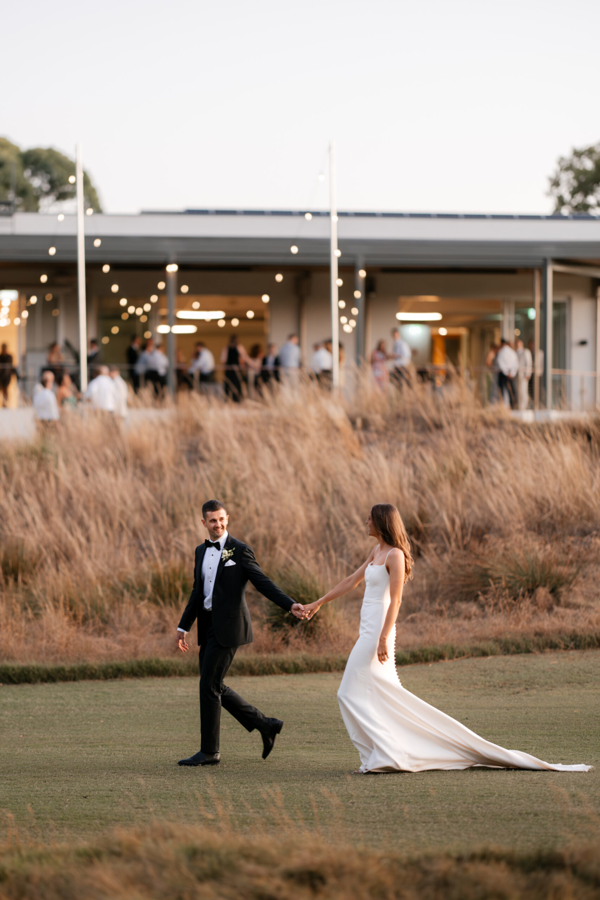 Bride and groom walk hand in hand on lawn in front of a modern venue with guests and string lights at dusk.