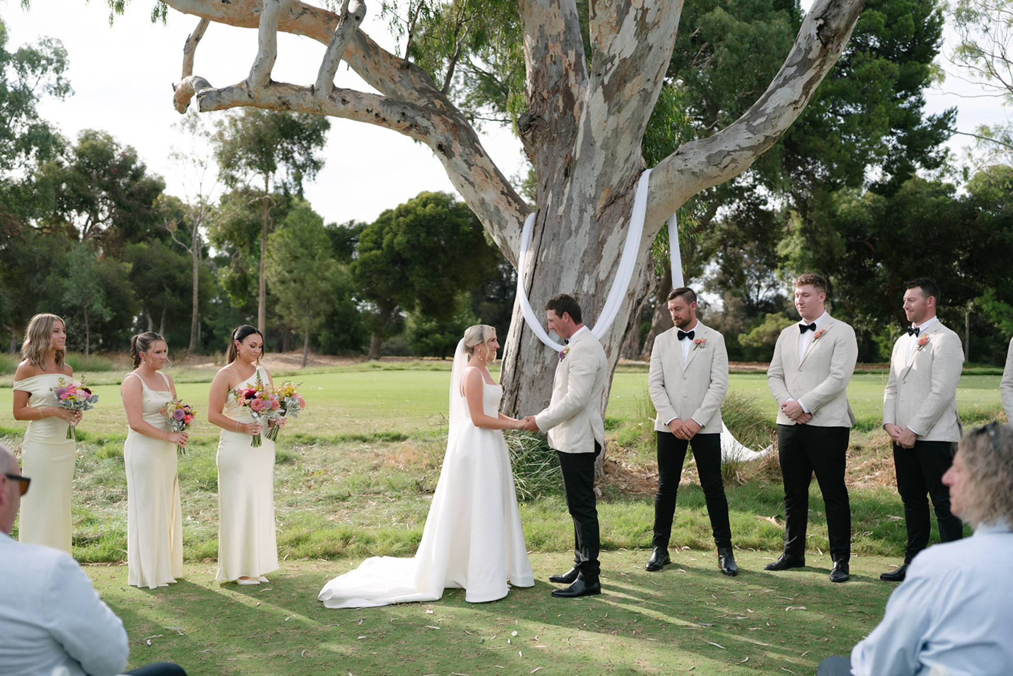 Outdoor wedding ceremony under a large tree with the couple and wedding party standing on a grassy lawn.