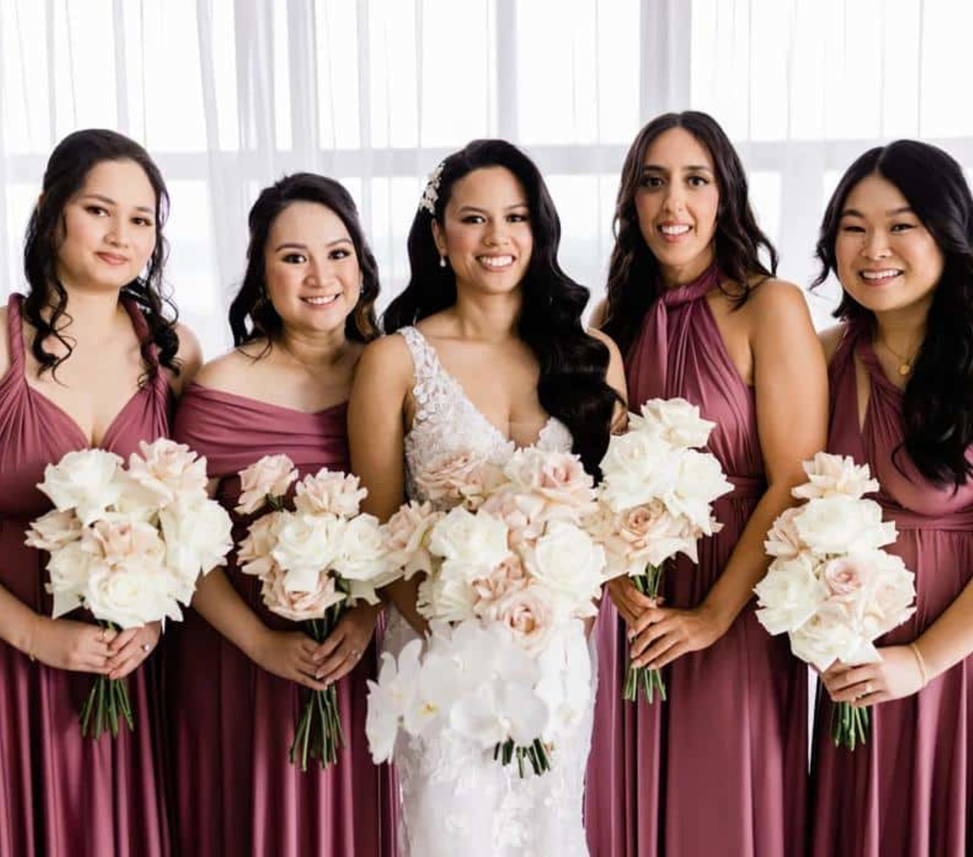 Bride and bridesmaids in mauve dresses holding blush and white rose bouquets in front of a bright window.