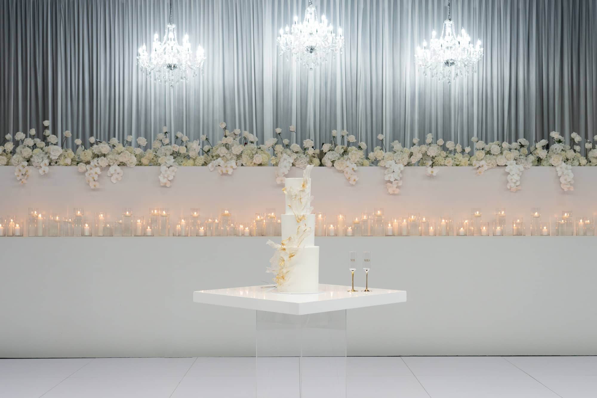 Tall white wedding cake on a modern table framed by candles, white florals, and crystal chandeliers.