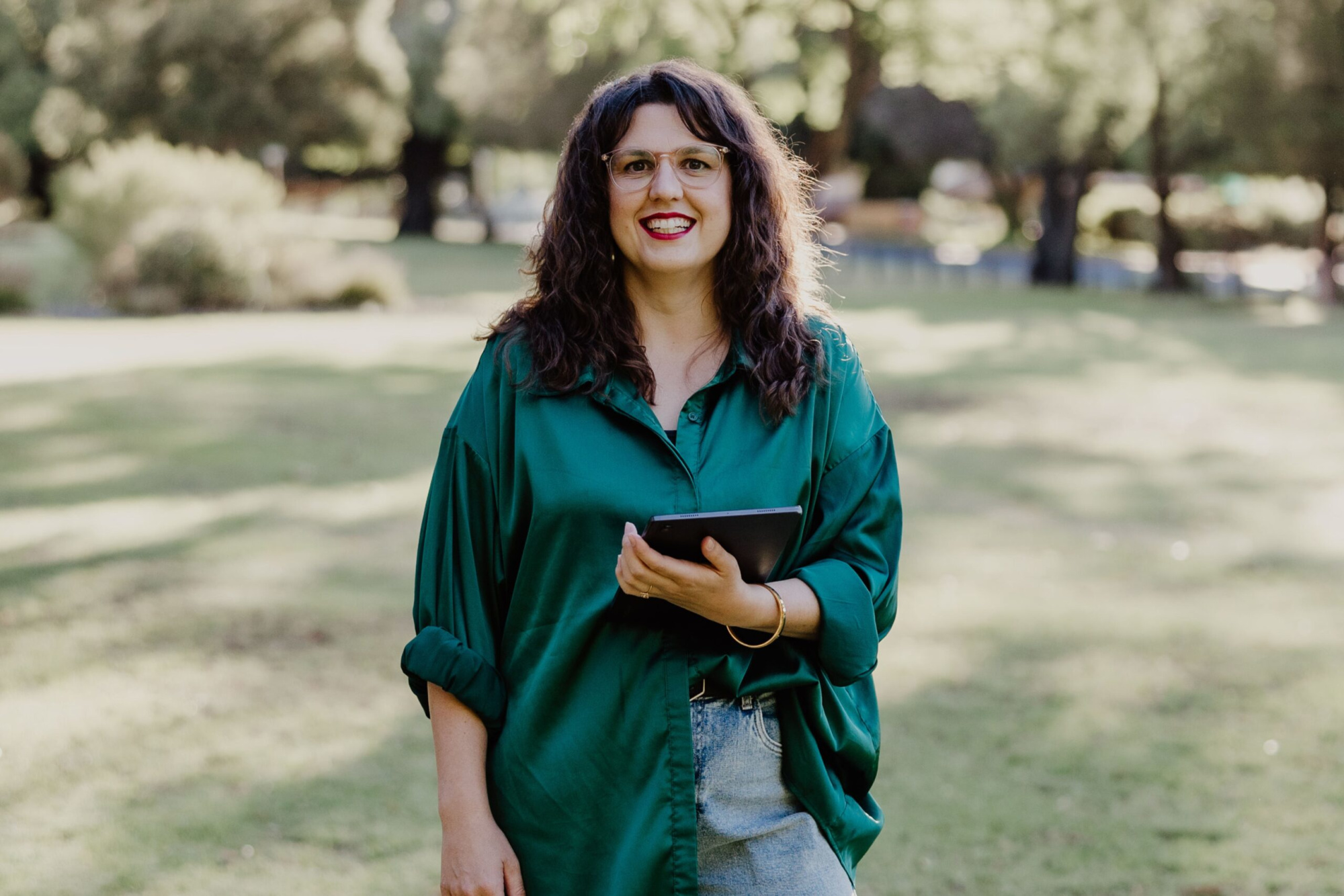 Smiling wedding planner holding a tablet while standing in a sunny outdoor park.