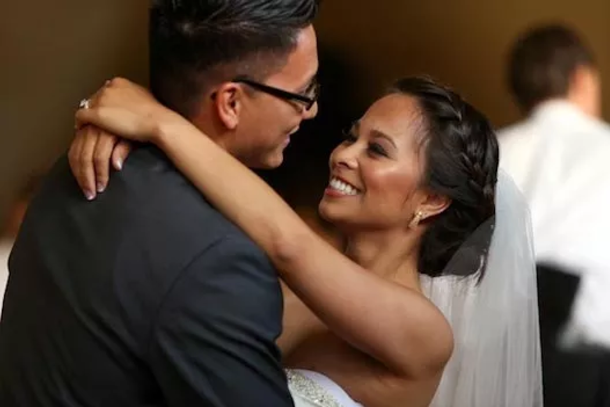 Bride and groom smile and embrace during their first dance at an indoor wedding reception.