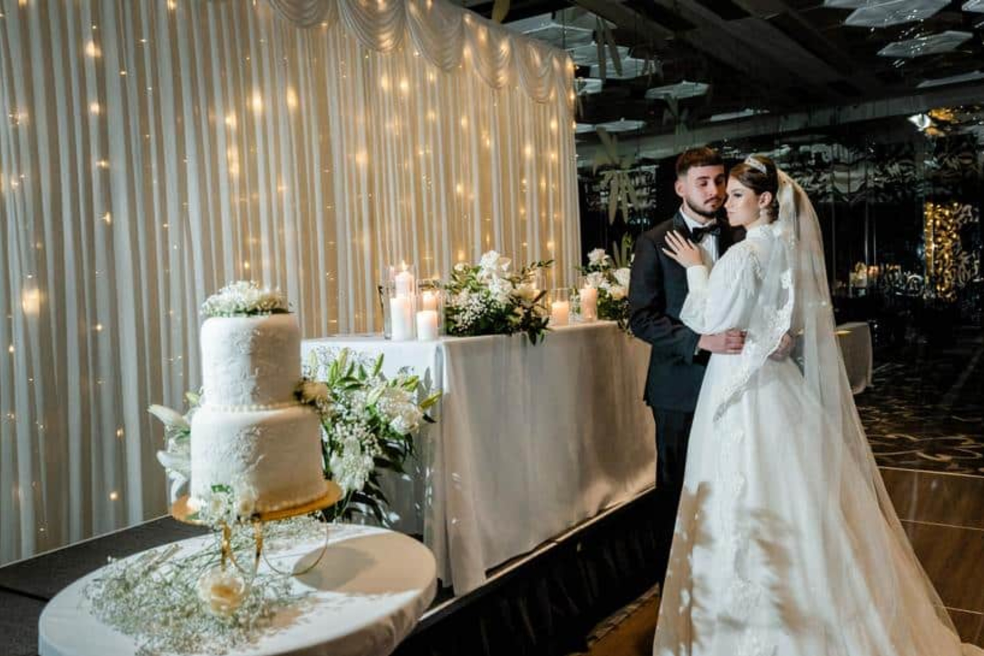 Bride and groom embrace beside a lit reception backdrop with candles, flowers, and a tiered white wedding cake.