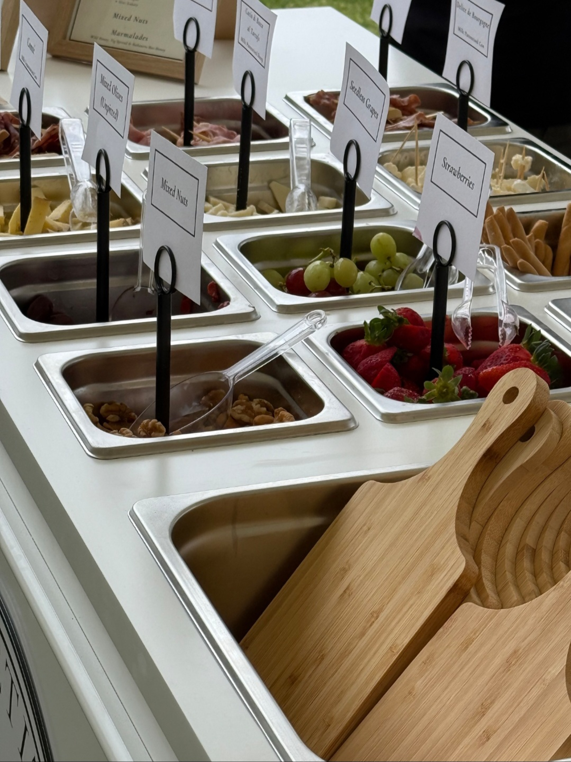Self-serve wedding grazing station with labeled trays of fruit, nuts, and snacks beside stacked wooden serving boards.