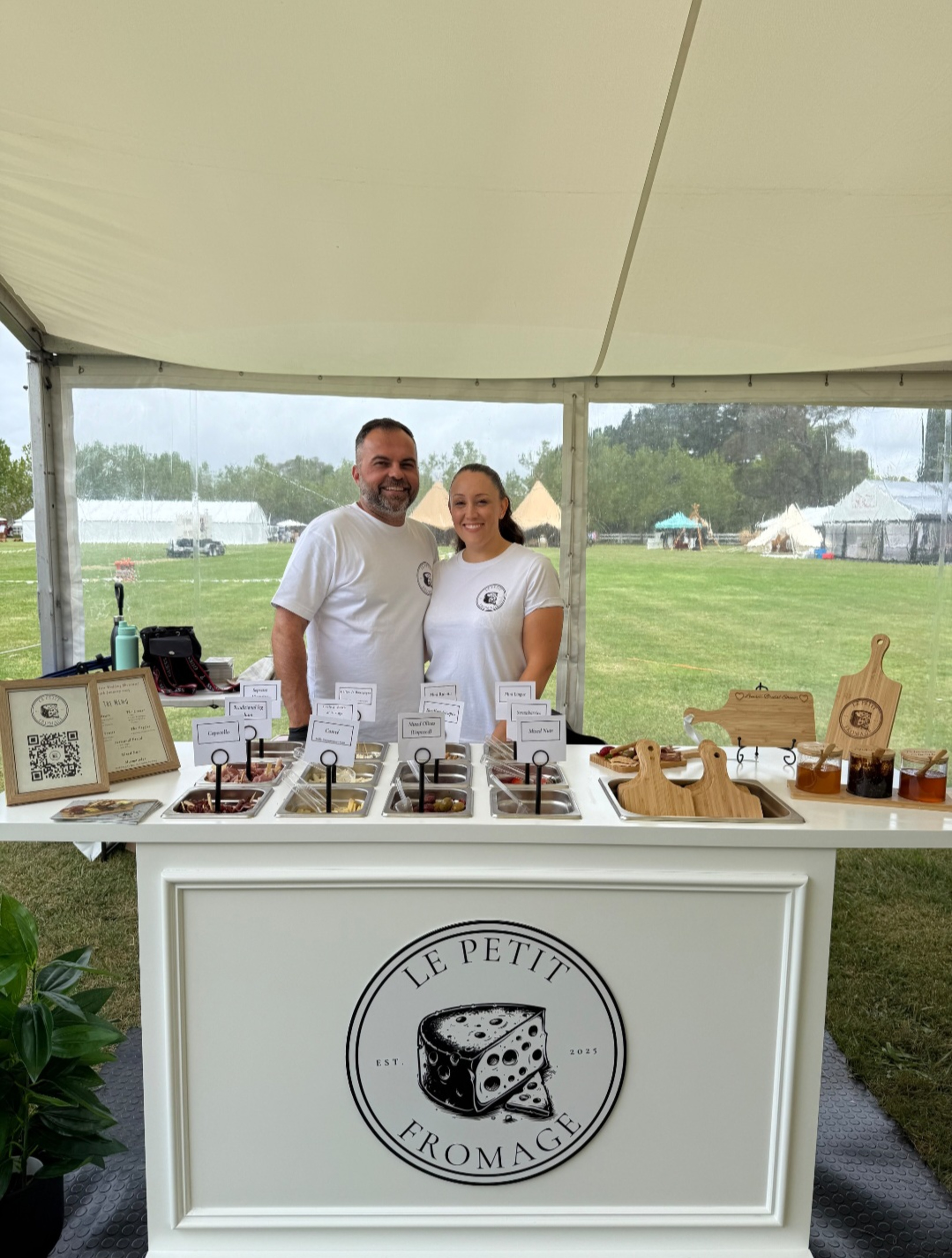 Smiling caterers stand behind a Le Petit Fromage cheese tasting station under an outdoor wedding tent.