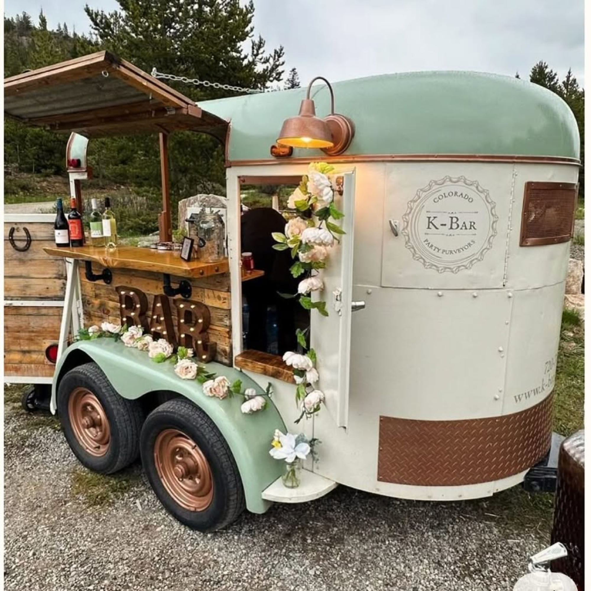 Rustic mobile bar trailer decorated with flowers and serving drinks at an outdoor wedding venue.