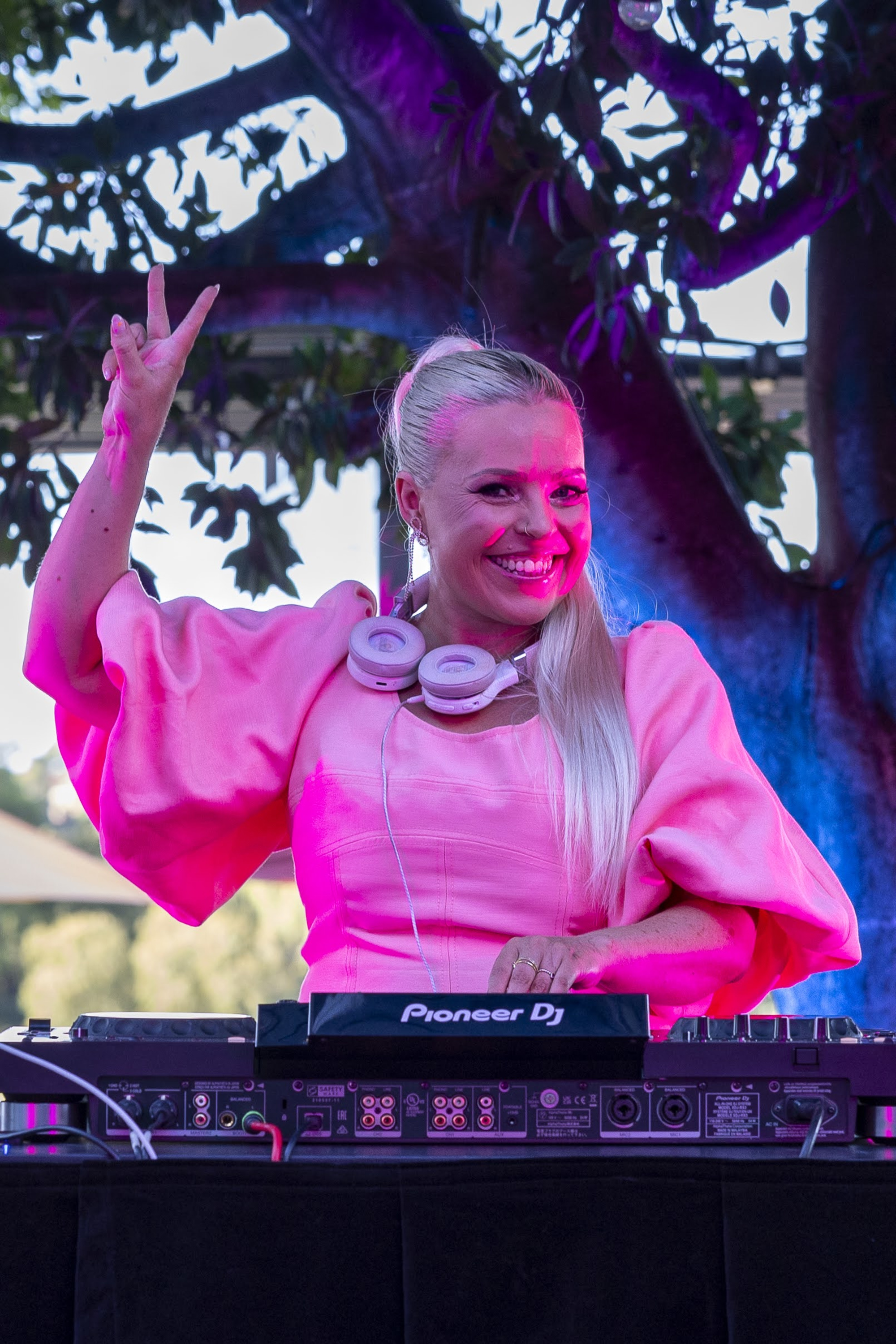 Smiling wedding DJ in a bright pink dress mixing music at an outdoor reception.