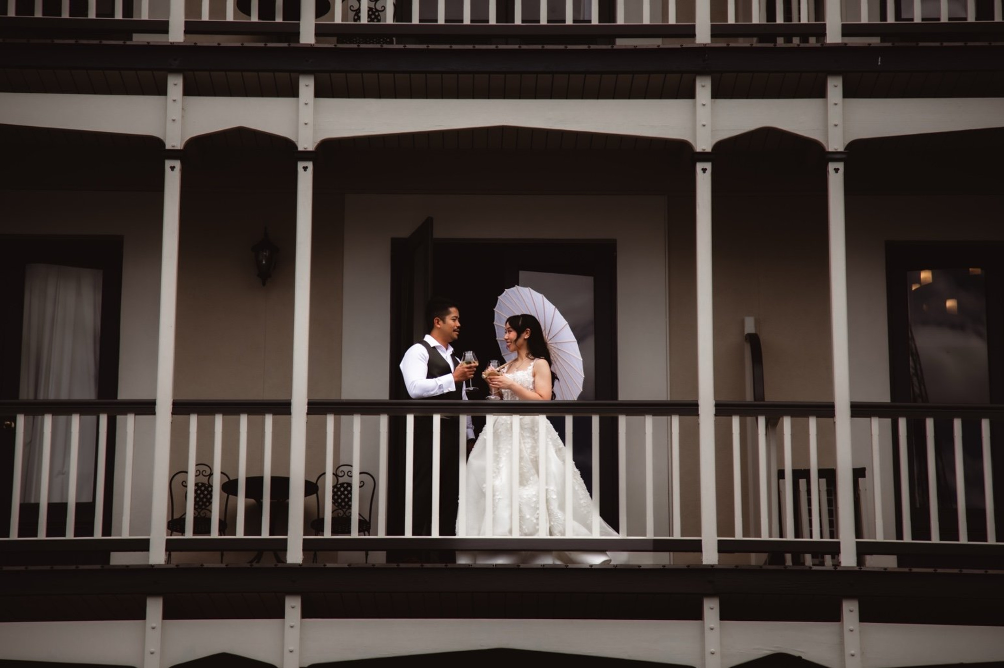 Bride and groom share a toast on a vintage balcony, with the bride holding a white parasol.