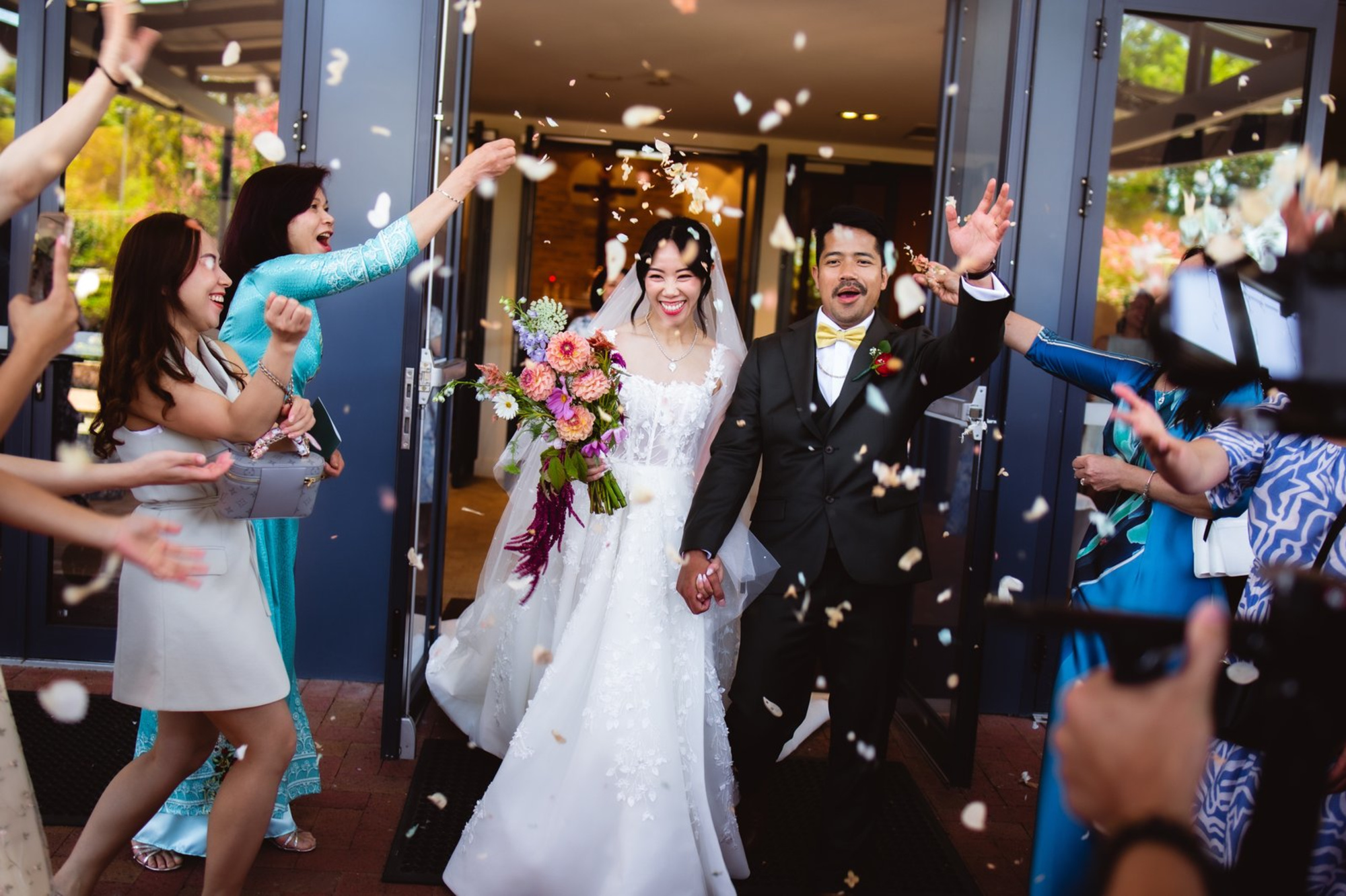 Bride and groom walk out of the venue smiling as guests toss flower petals in celebration.