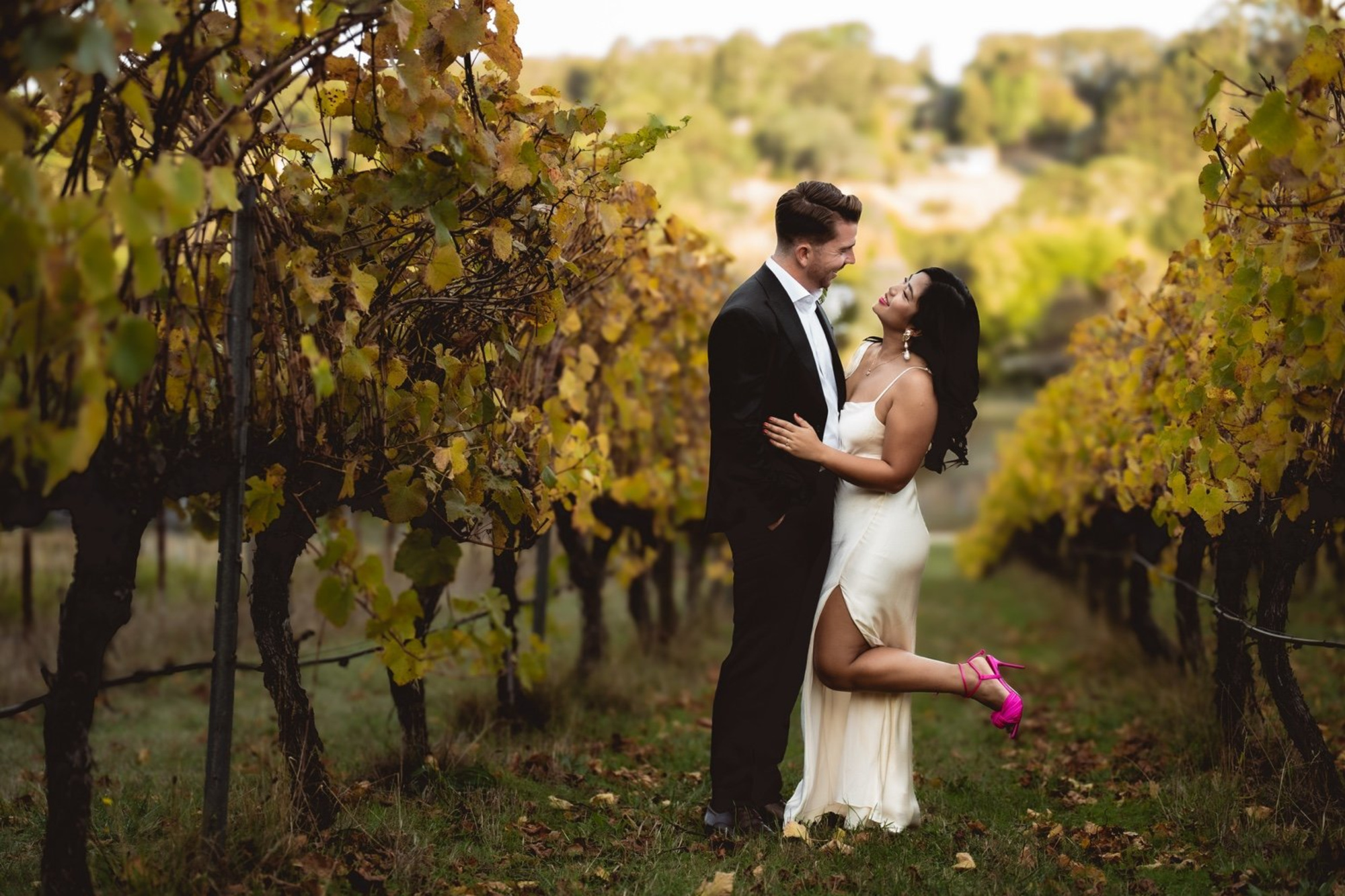 A couple in formal wedding attire embrace in a vineyard lined with golden leaves.