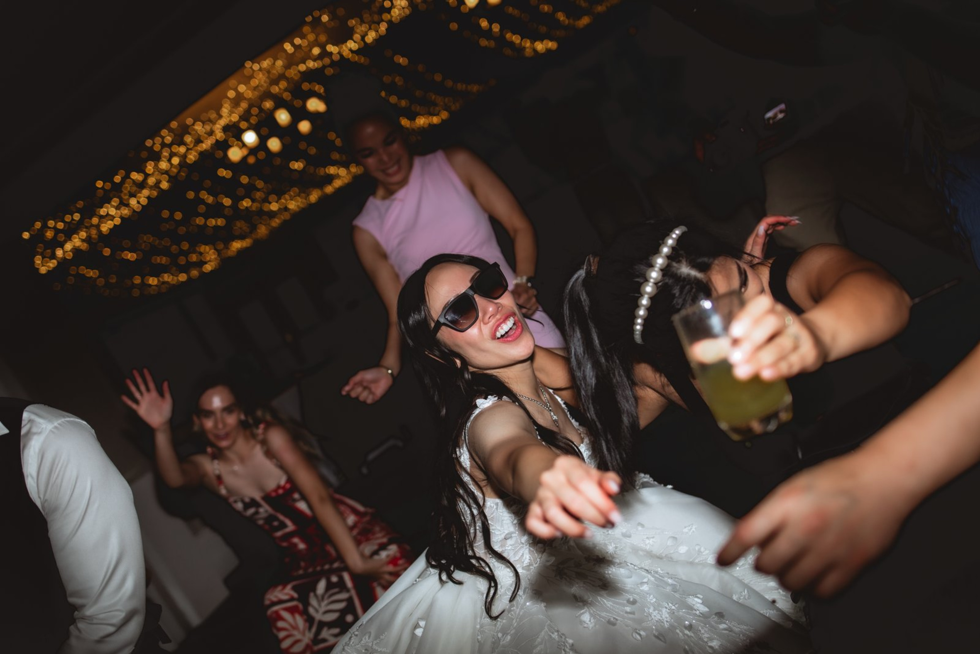 Bride in sunglasses dances with friends under warm string lights at a lively wedding reception.