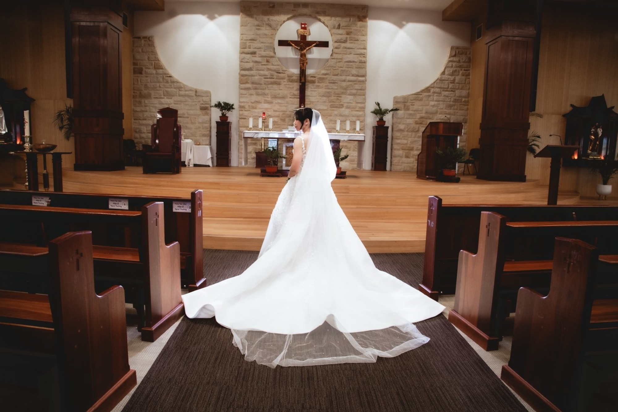 Bride in a long white gown stands facing the altar in a church with wooden pews and stone backdrop.