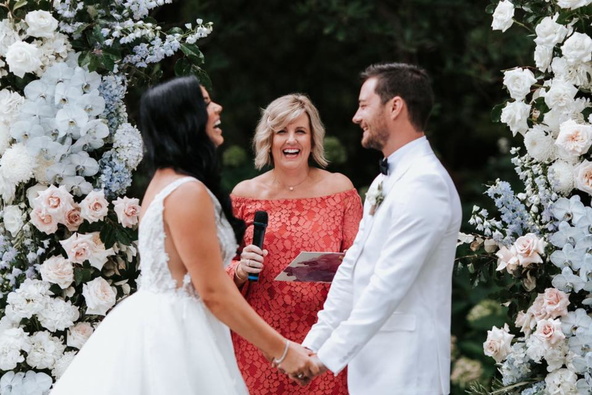 Bride and groom laughing while holding hands under a lush floral arch with an officiant during an outdoor wedding ceremony.