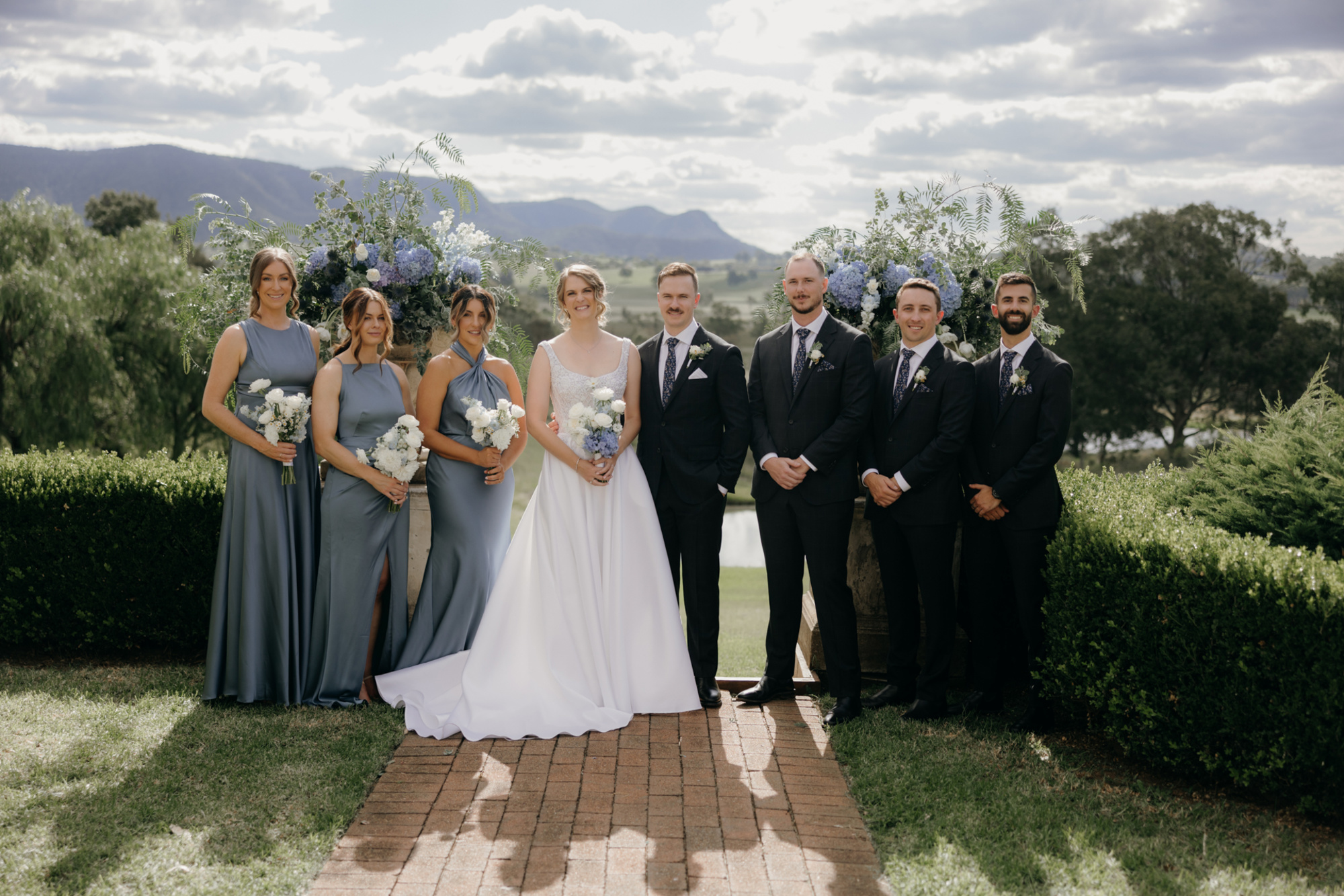 Bride and groom posing with bridesmaids and groomsmen in blue tones at an outdoor garden wedding with mountain views.