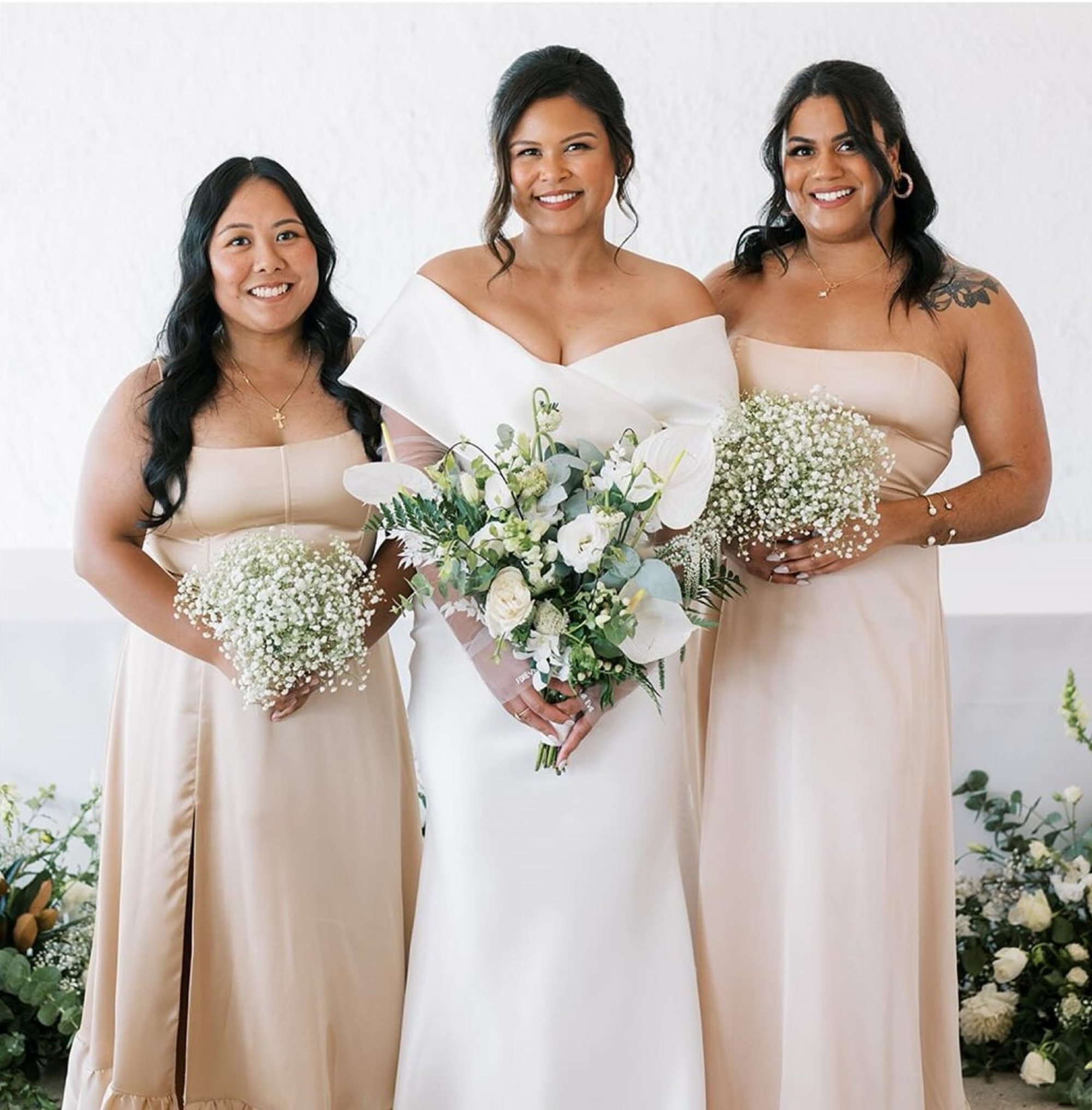 Bride in a white gown stands between two bridesmaids in champagne dresses holding white floral bouquets.
