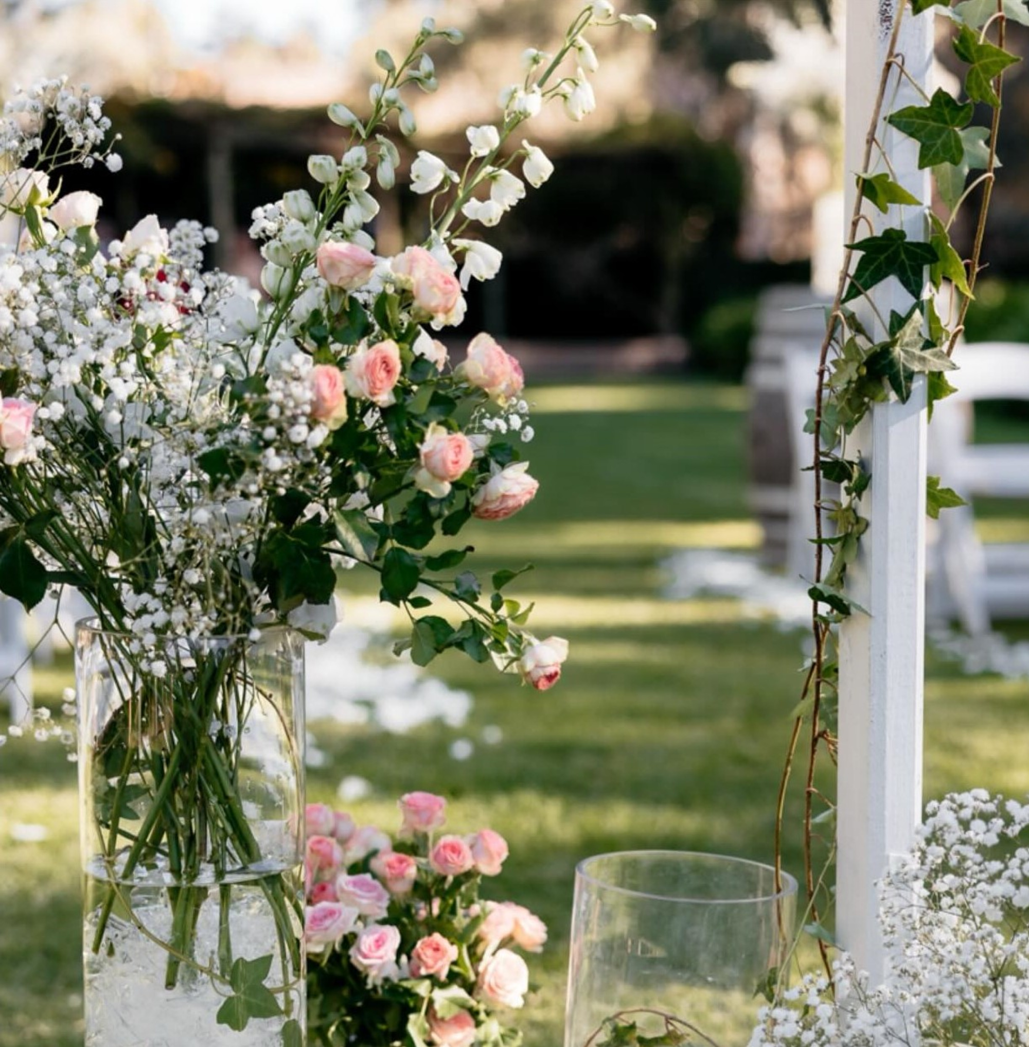 Outdoor wedding ceremony decor with pink roses and white flowers arranged in tall glass vases by the aisle.