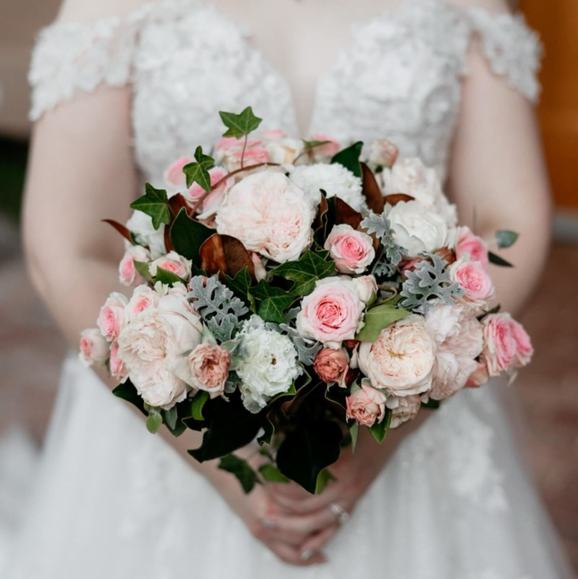 Bride in lace gown holding a lush pink and white rose bouquet with greenery and soft foliage.