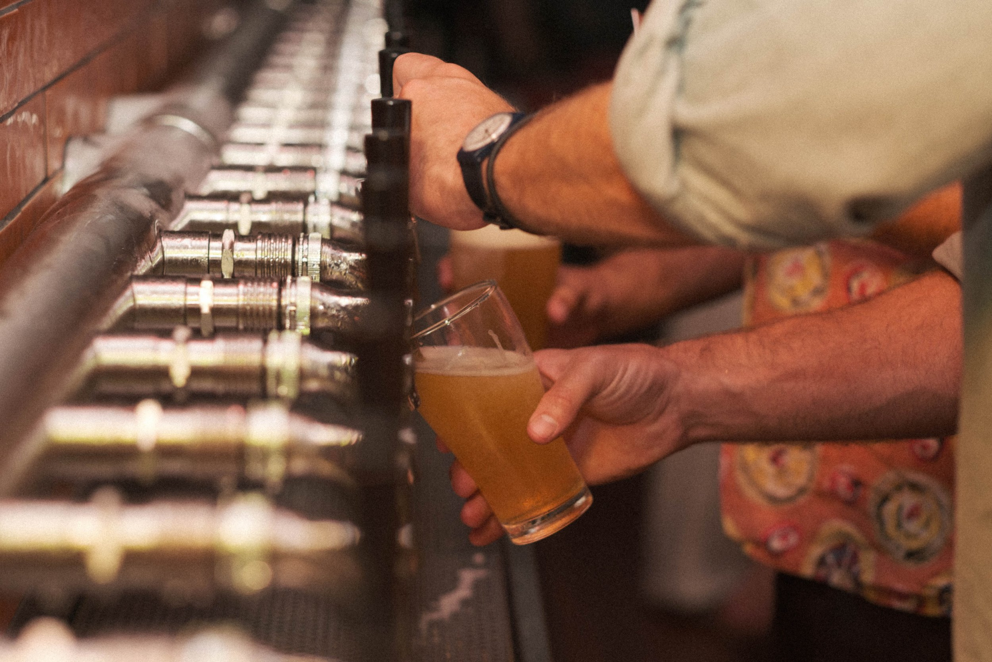 Guests pour draft beer from a long row of bar taps into pint glasses at a wedding reception bar.