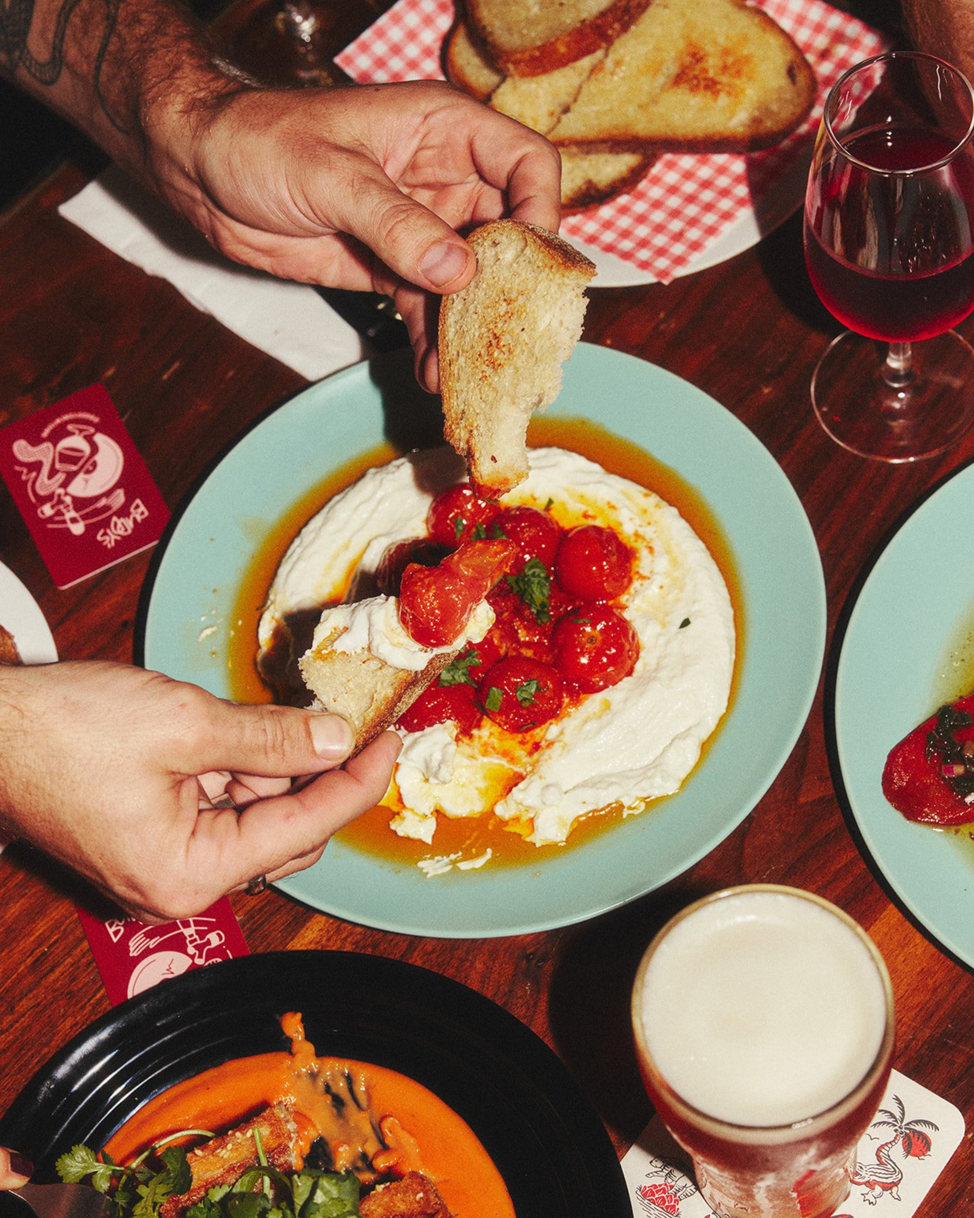 Hands dip toasted bread into a plate of creamy cheese topped with tomatoes at a wedding-style shared appetizer table.