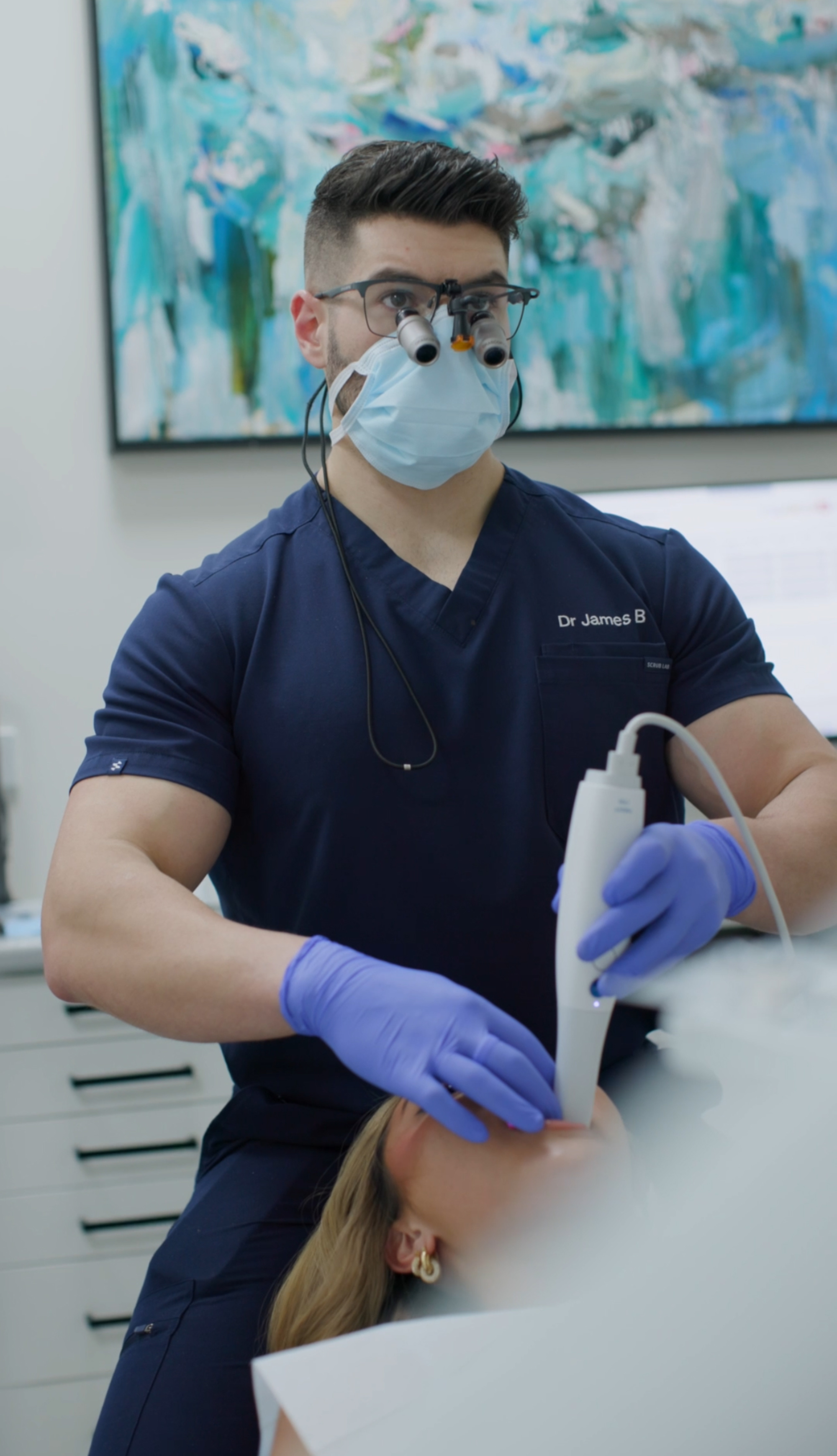 A clinician in scrubs and mask performs a facial skincare treatment on a woman lying in a clinic chair.