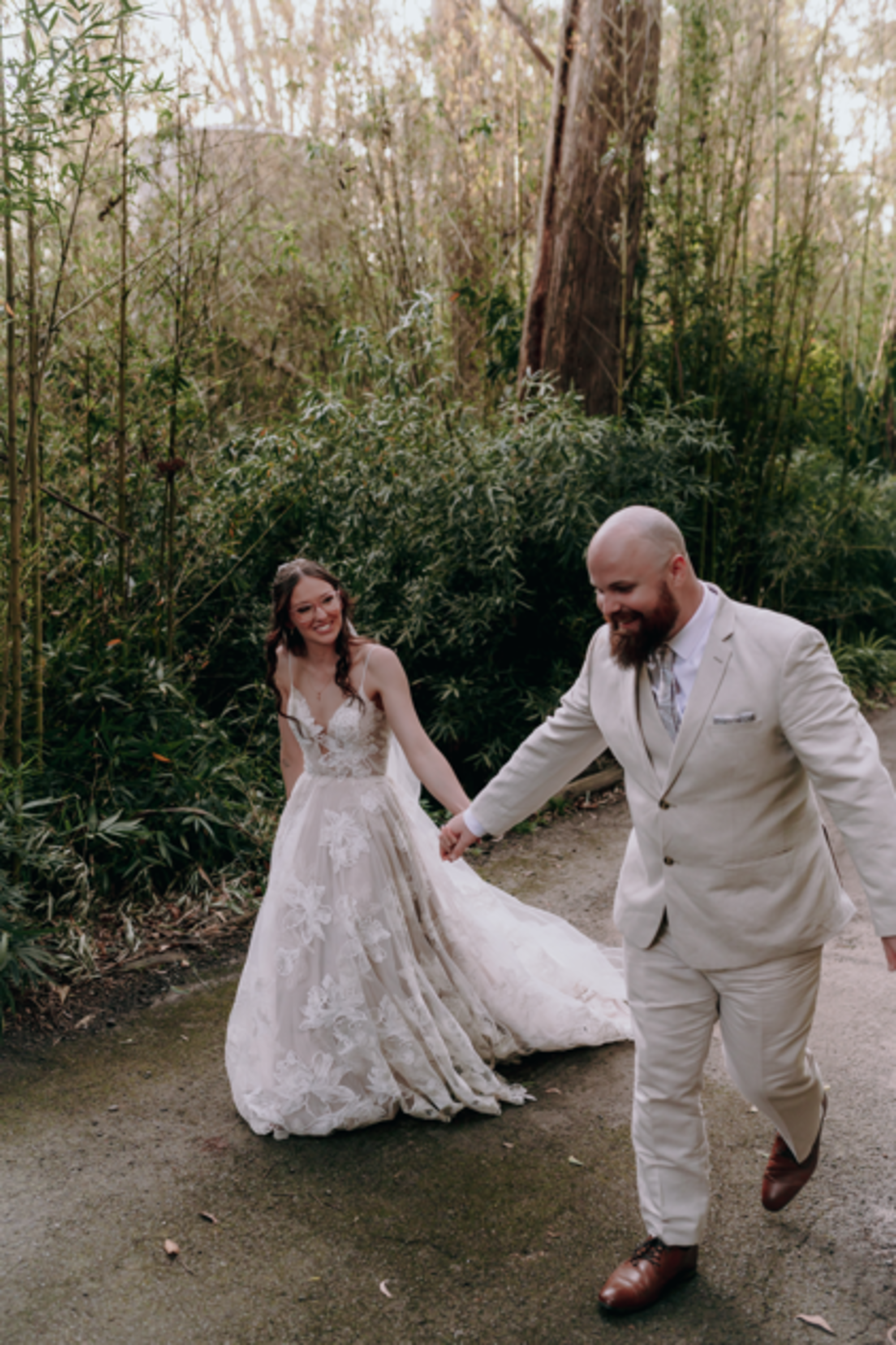 Bride and groom walk hand in hand along a forest path, laughing together in their wedding attire.