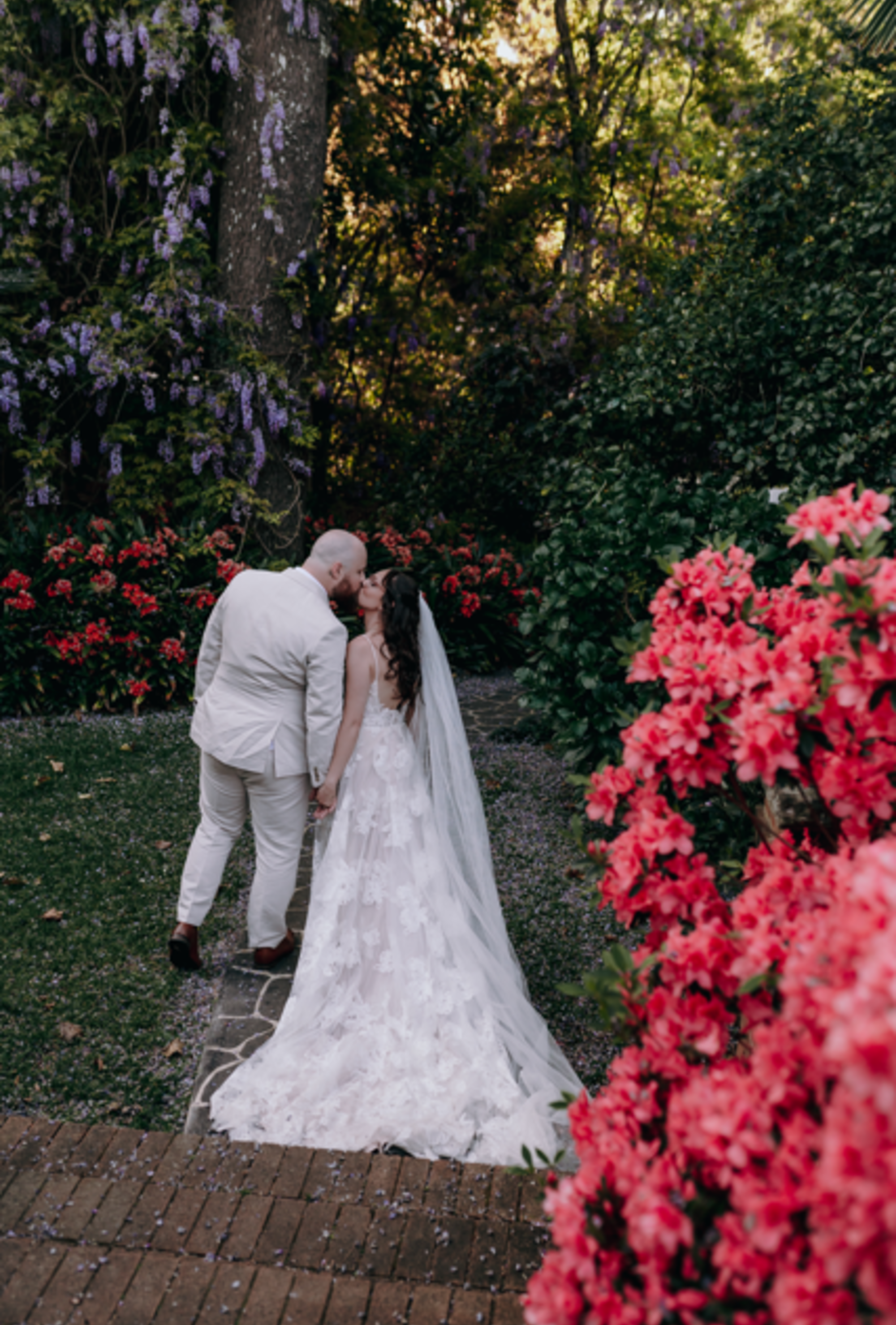 A bride and groom share a kiss in a lush garden pathway surrounded by vibrant flowers and greenery.
