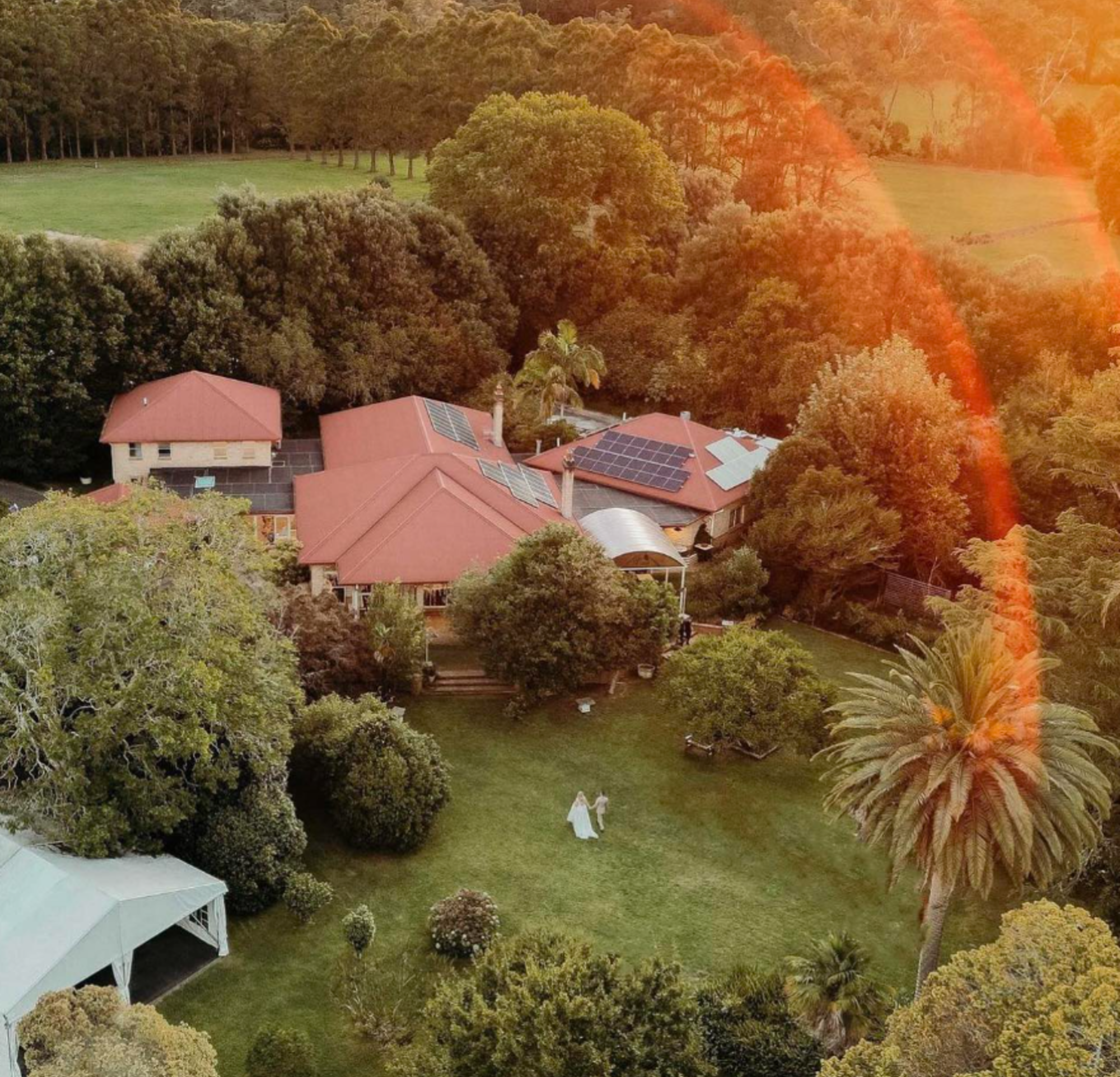Aerial view of a countryside estate wedding venue with lush gardens and a couple walking on the lawn at sunset.