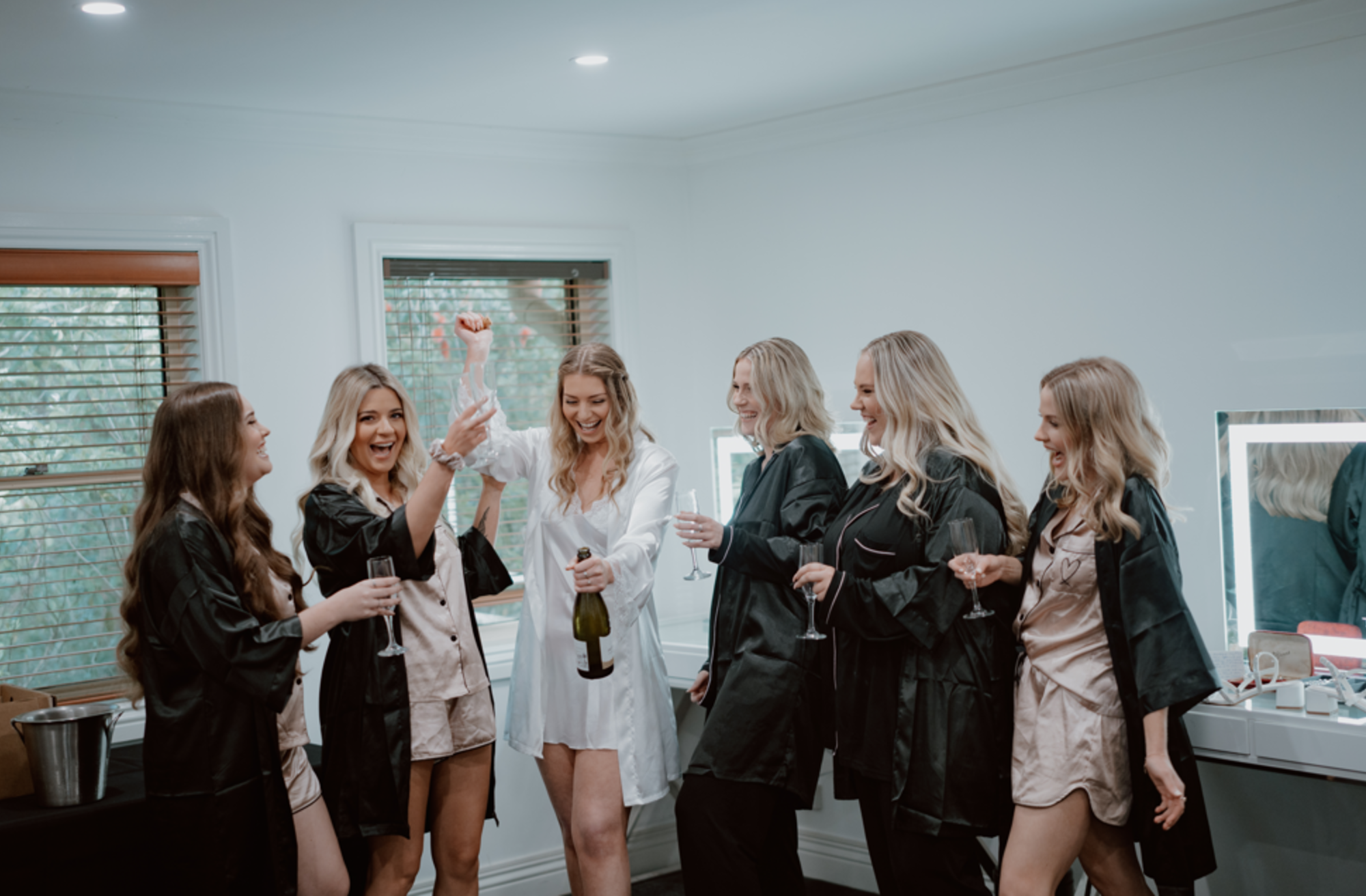 Bride and bridesmaids in satin robes celebrate with champagne in a bright bridal suite.