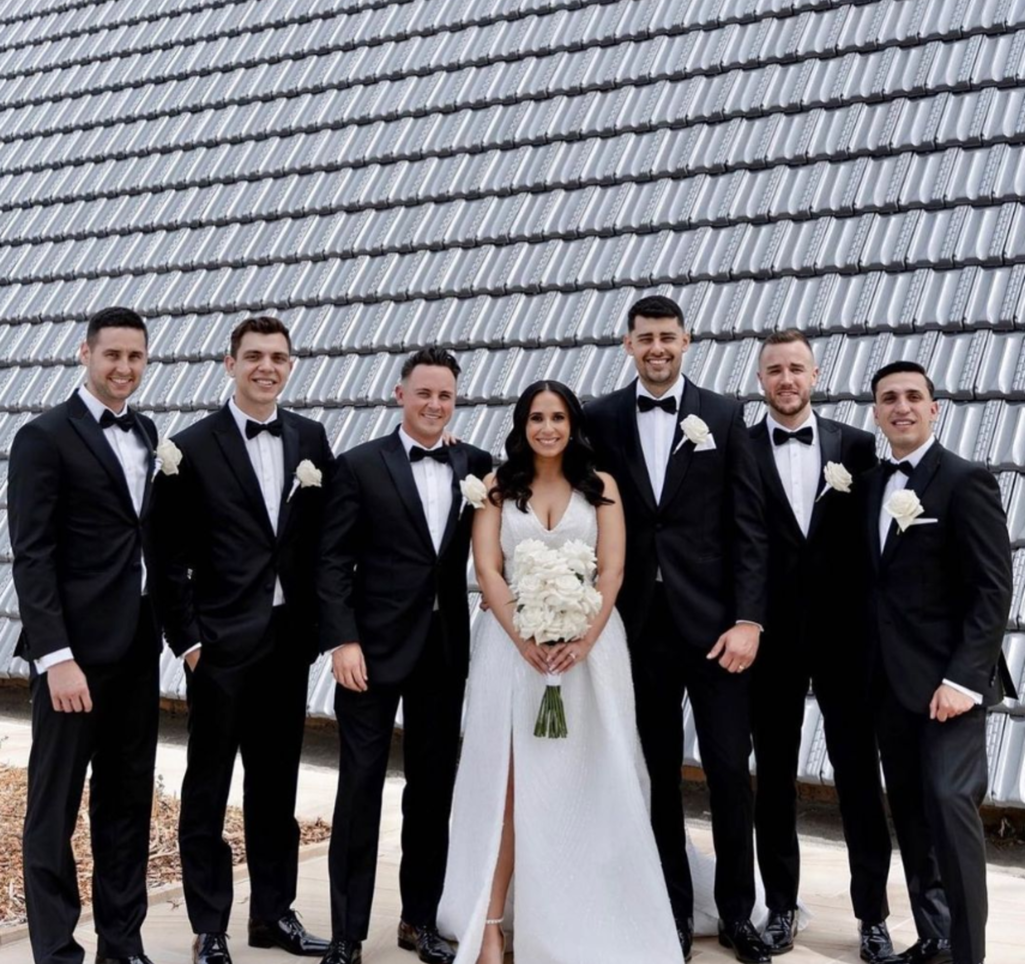 Bride in white gown poses with groomsmen in black tuxedos against a modern tiled wall backdrop.