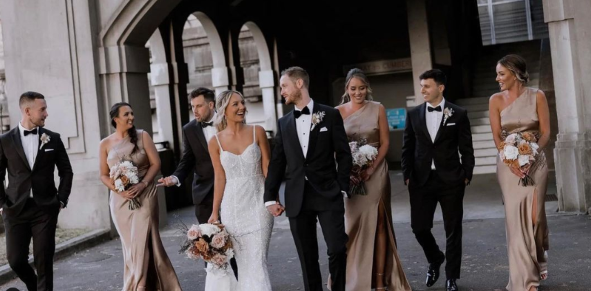Bride and groom walk with their wedding party in elegant attire under an urban archway.