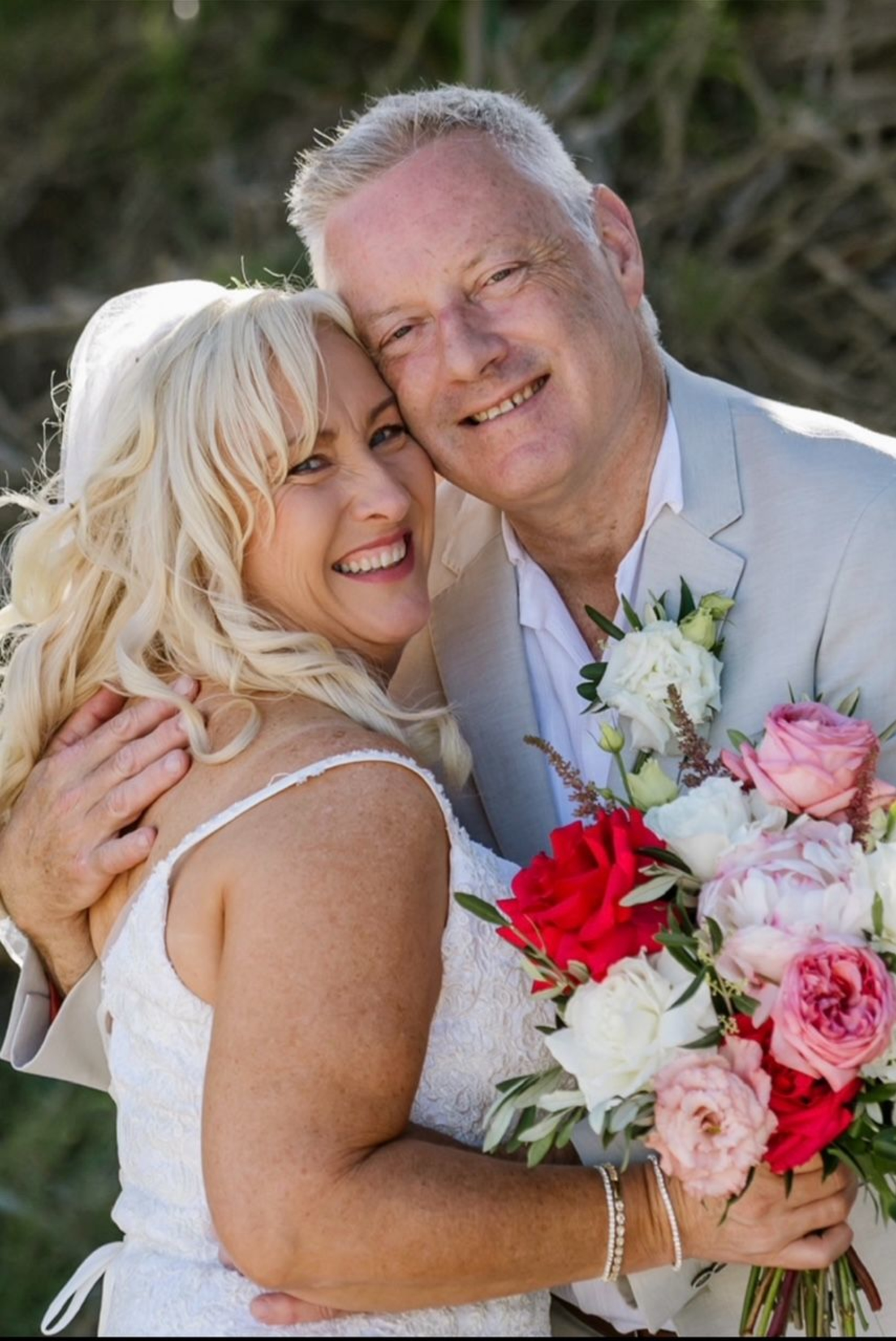 Smiling bride and groom embrace outdoors, holding a vibrant bouquet of pink, red, and white flowers.
