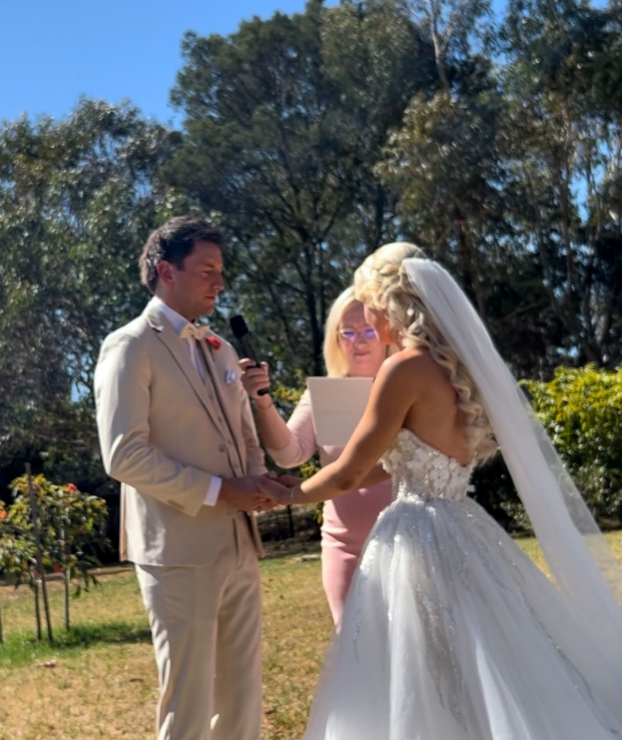 Bride and groom hold hands during an outdoor wedding ceremony with an officiant reading vows.