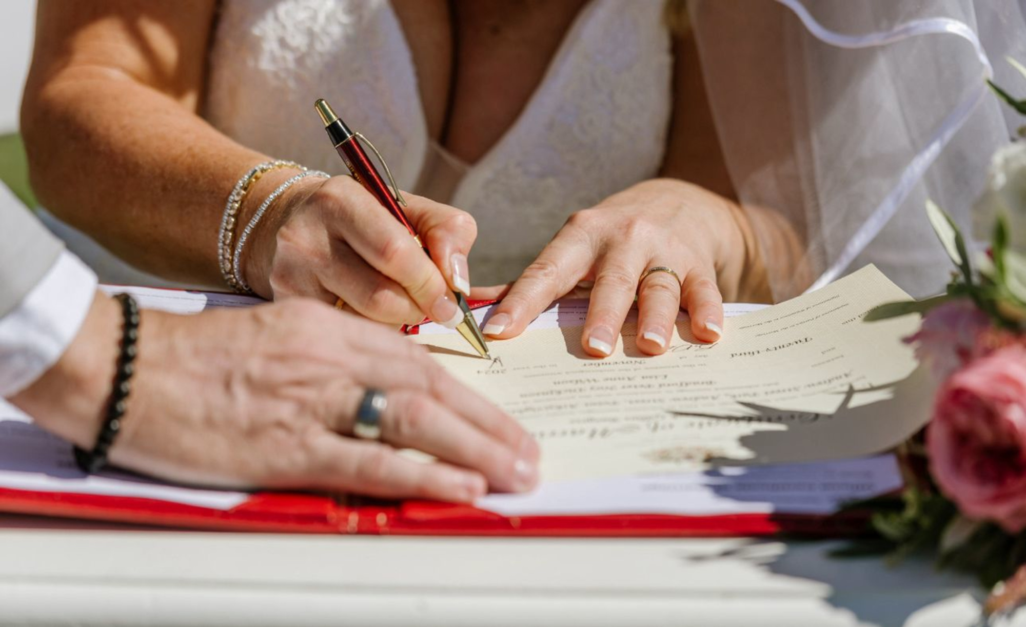 Close-up of a bride signing the marriage certificate beside the groom during an outdoor wedding ceremony.