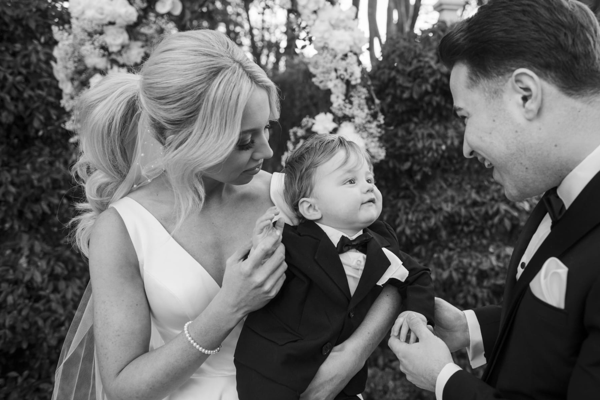 Bride and groom smile at their baby in a tiny tuxedo during an outdoor wedding portrait.