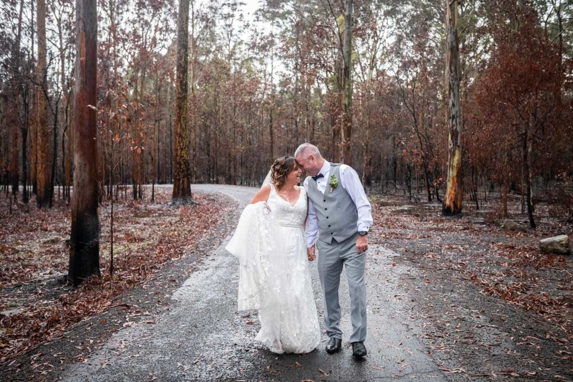 A bride and groom walk hand in hand down a wet forest road, smiling closely together among tall trees.