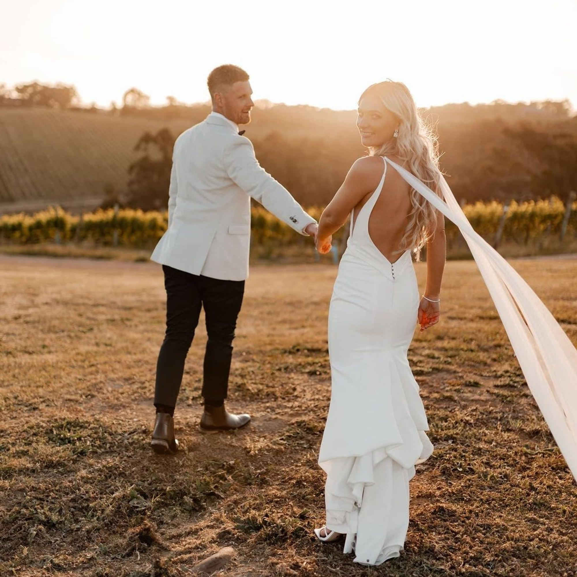 Bride and groom walk hand in hand through a sunlit vineyard field at sunset.