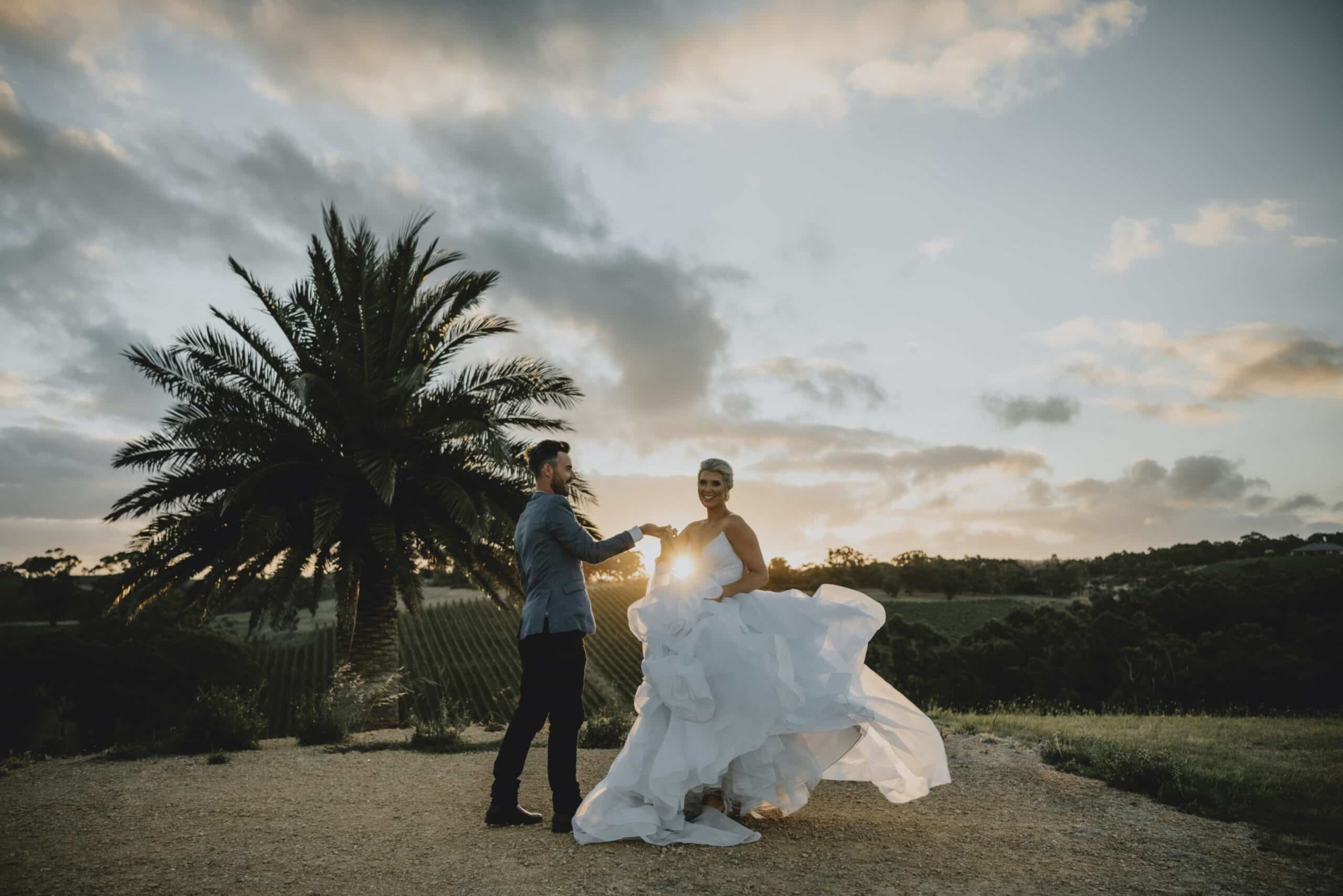 Bride and groom dance at sunset on a hilltop vineyard with a flowing dress and palm tree backdrop.